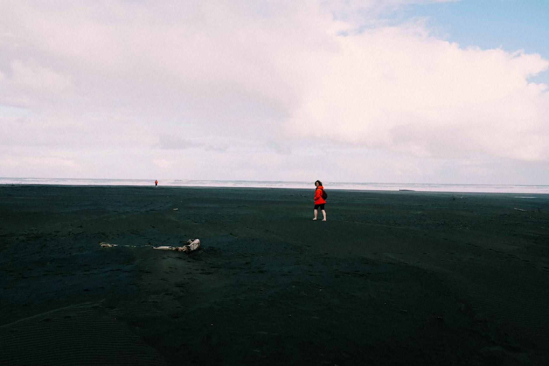 A solitary woman in a red jacket walks on a dark sandy beach under a cloudy sky.