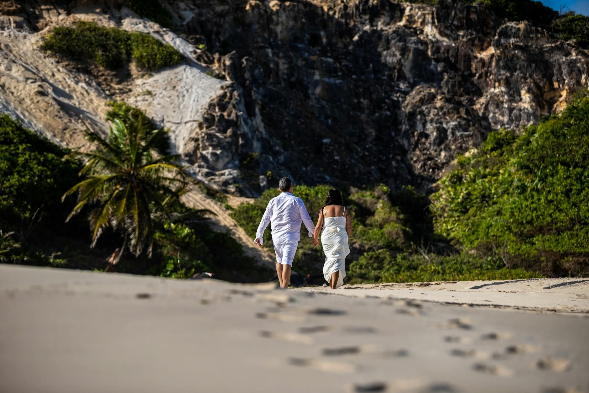 Romantic couple walking hand in hand on a scenic beach in PB, Brazil, with lush cliffs in the background.