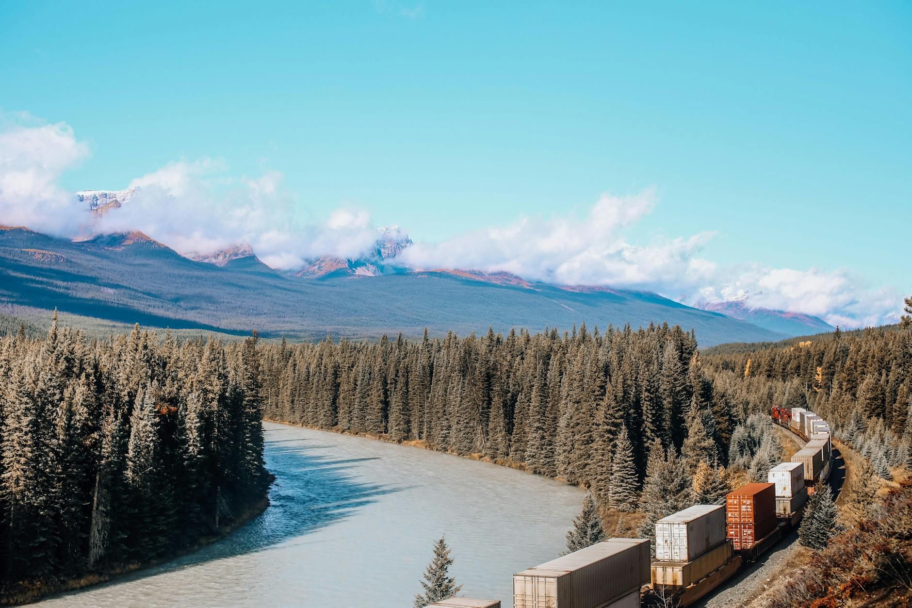 A cargo train travels alongside a lush river through a mountainous forest landscape on a clear day.