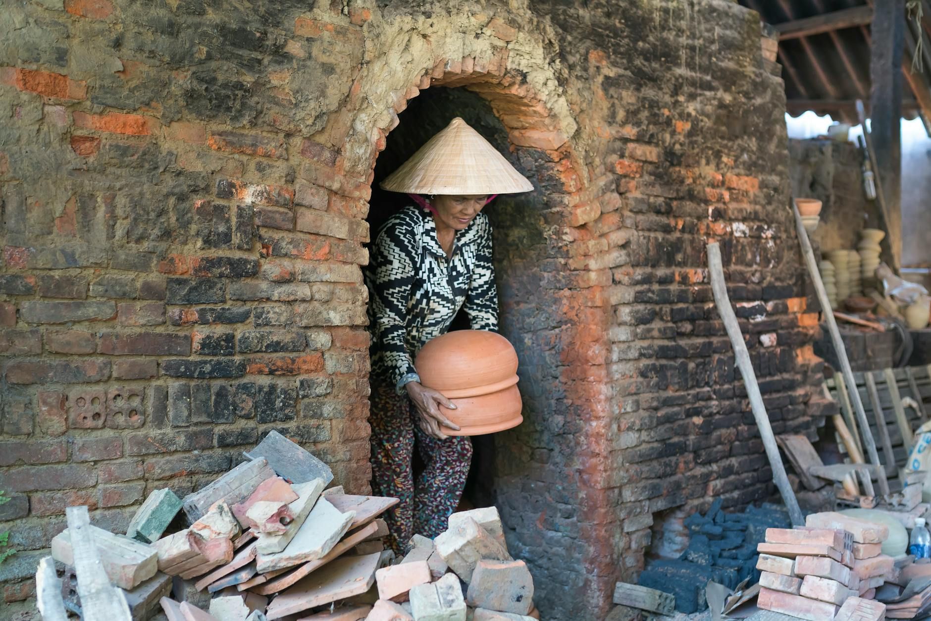 Woman in conical hat crafting clay pot in traditional brick kiln setting.