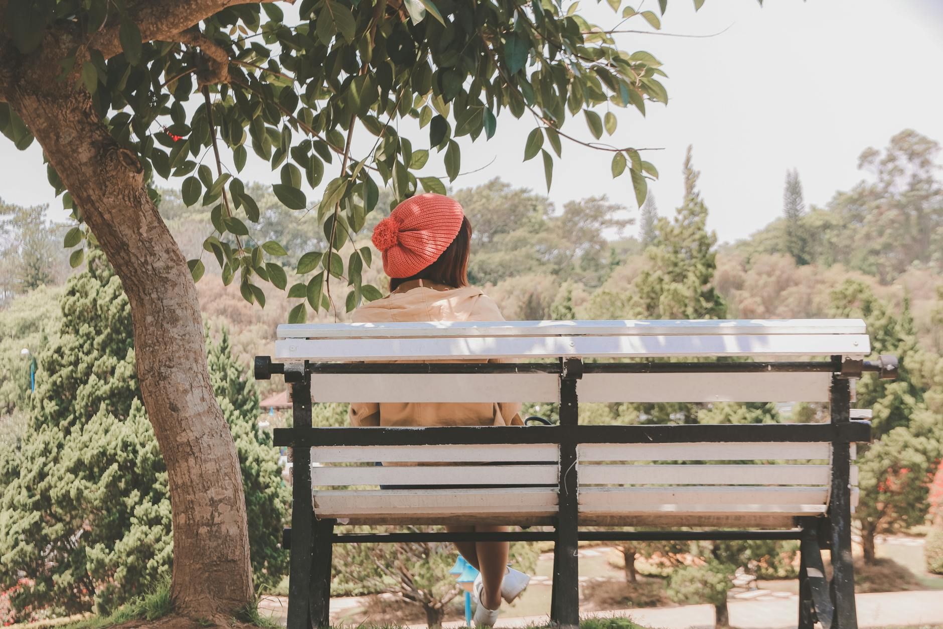 A woman wearing a red hat sits on a bench under a tree in a peaceful park.