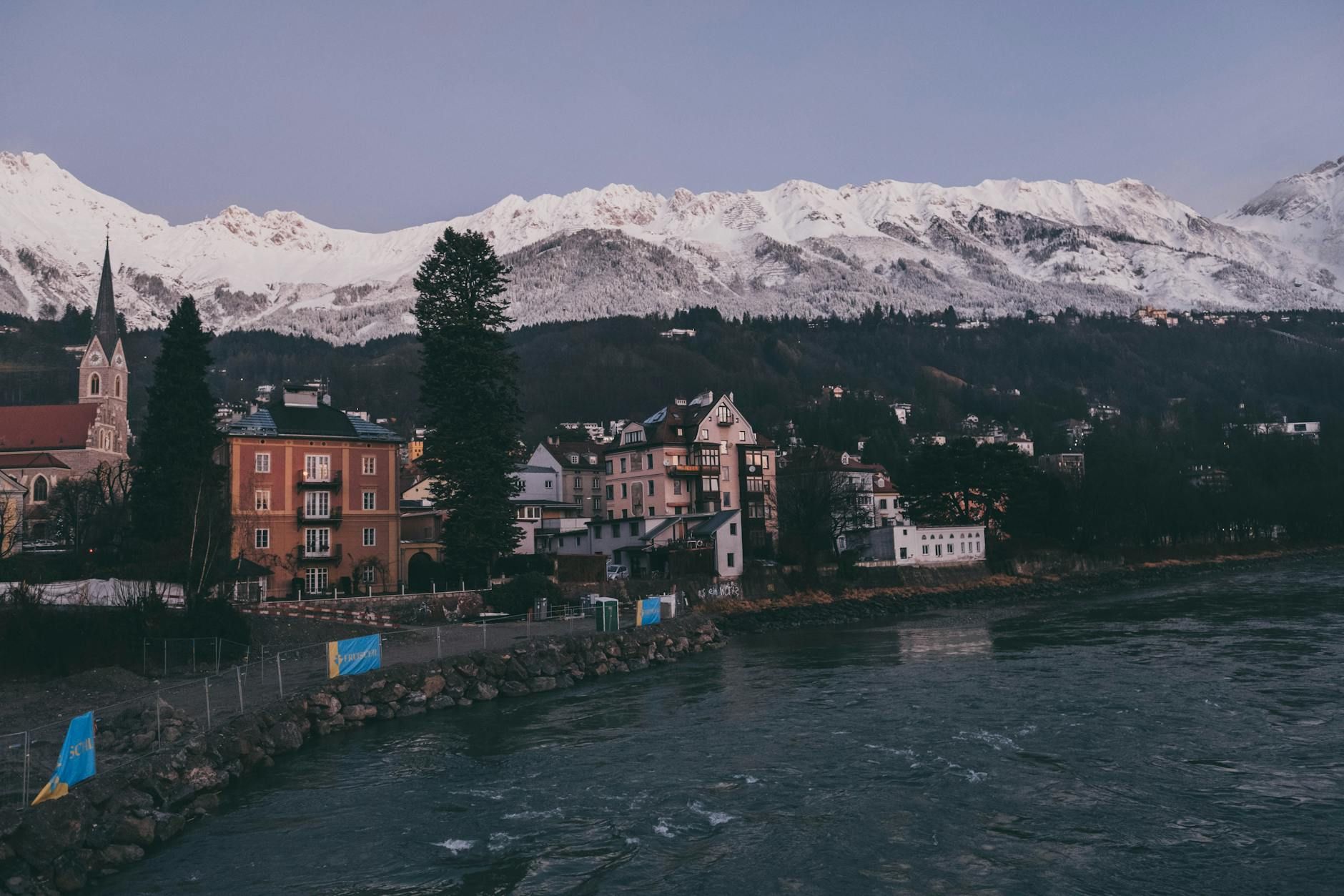 Charming Austrian town nestled by a river and snowcapped mountains at dusk.
