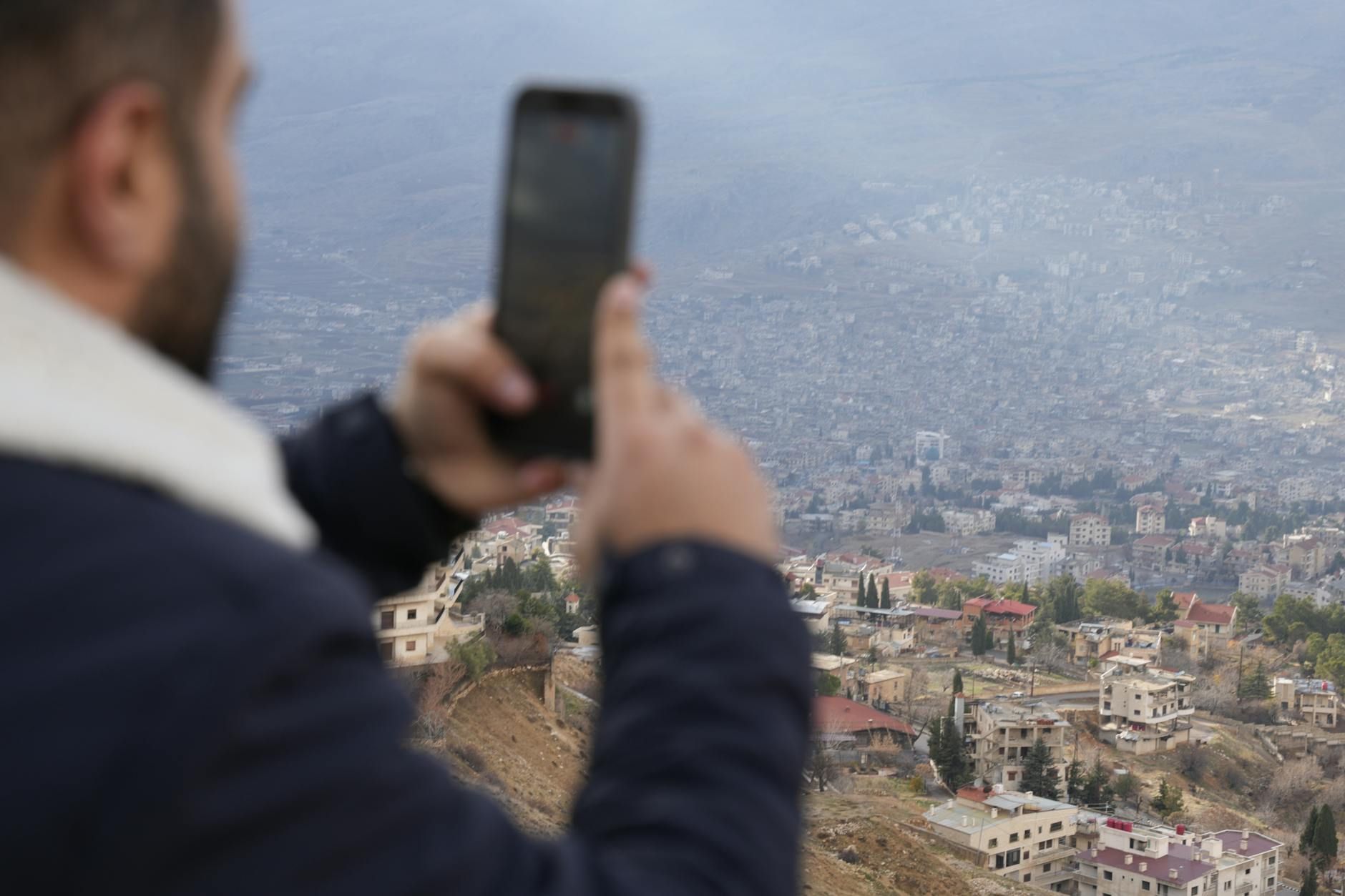 A person capturing a panoramic view of a Syrian cityscape from an elevated position with a smartphone.