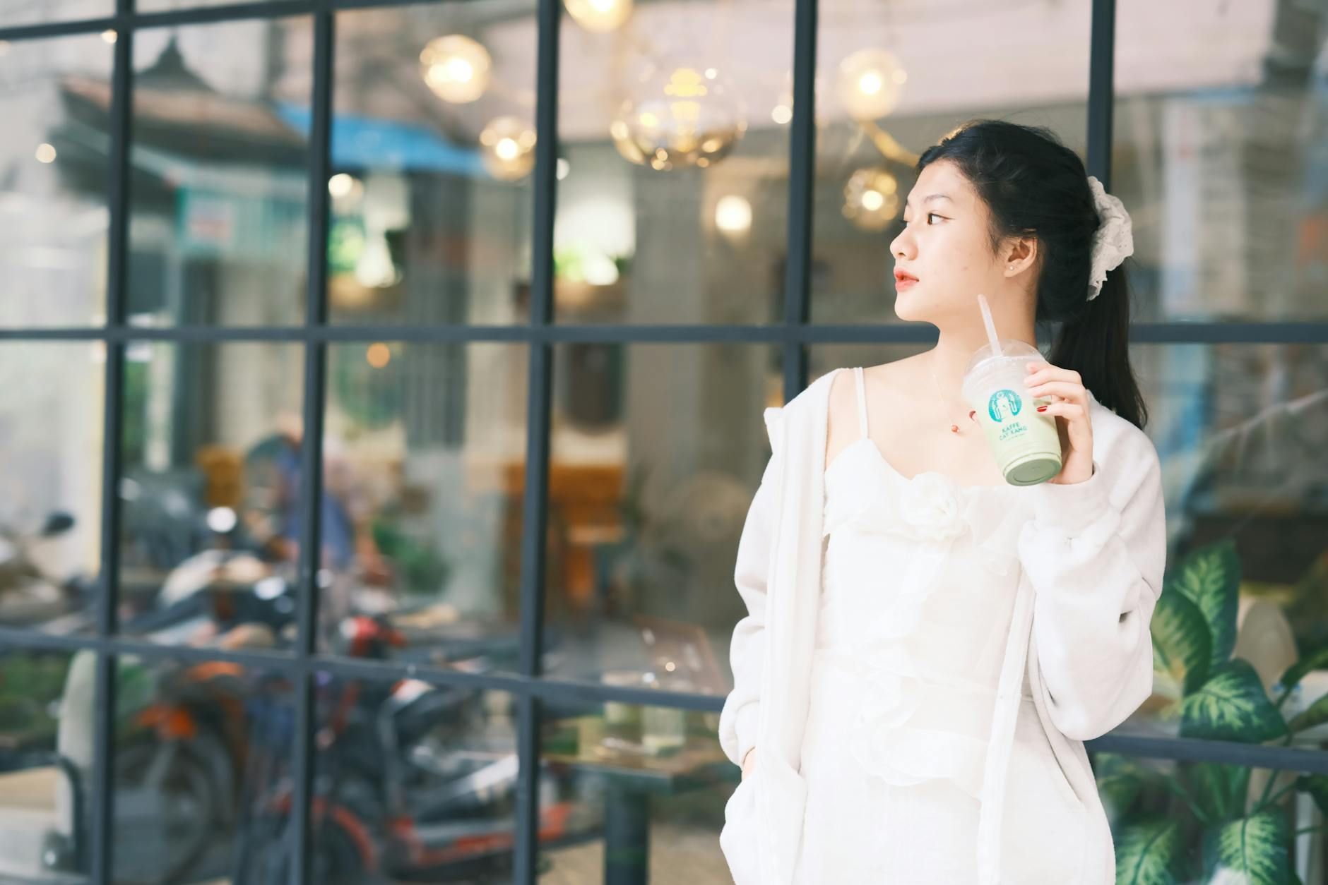 A young woman in white stands outside a cafe in Vietnam, holding a drink.