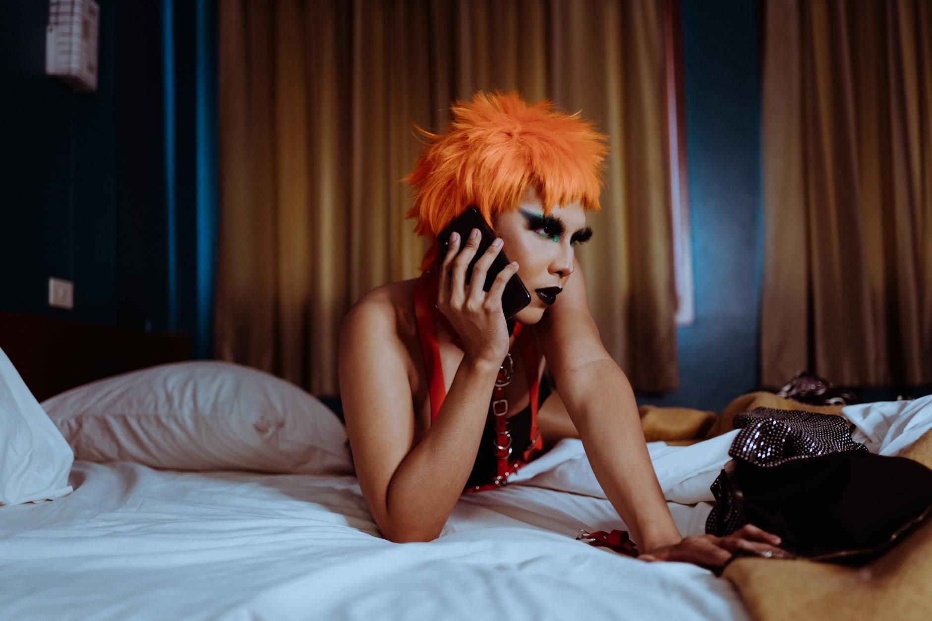 Seductive young ethnic female with makeup in orange wig lying on bed and chatting on mobile phone
