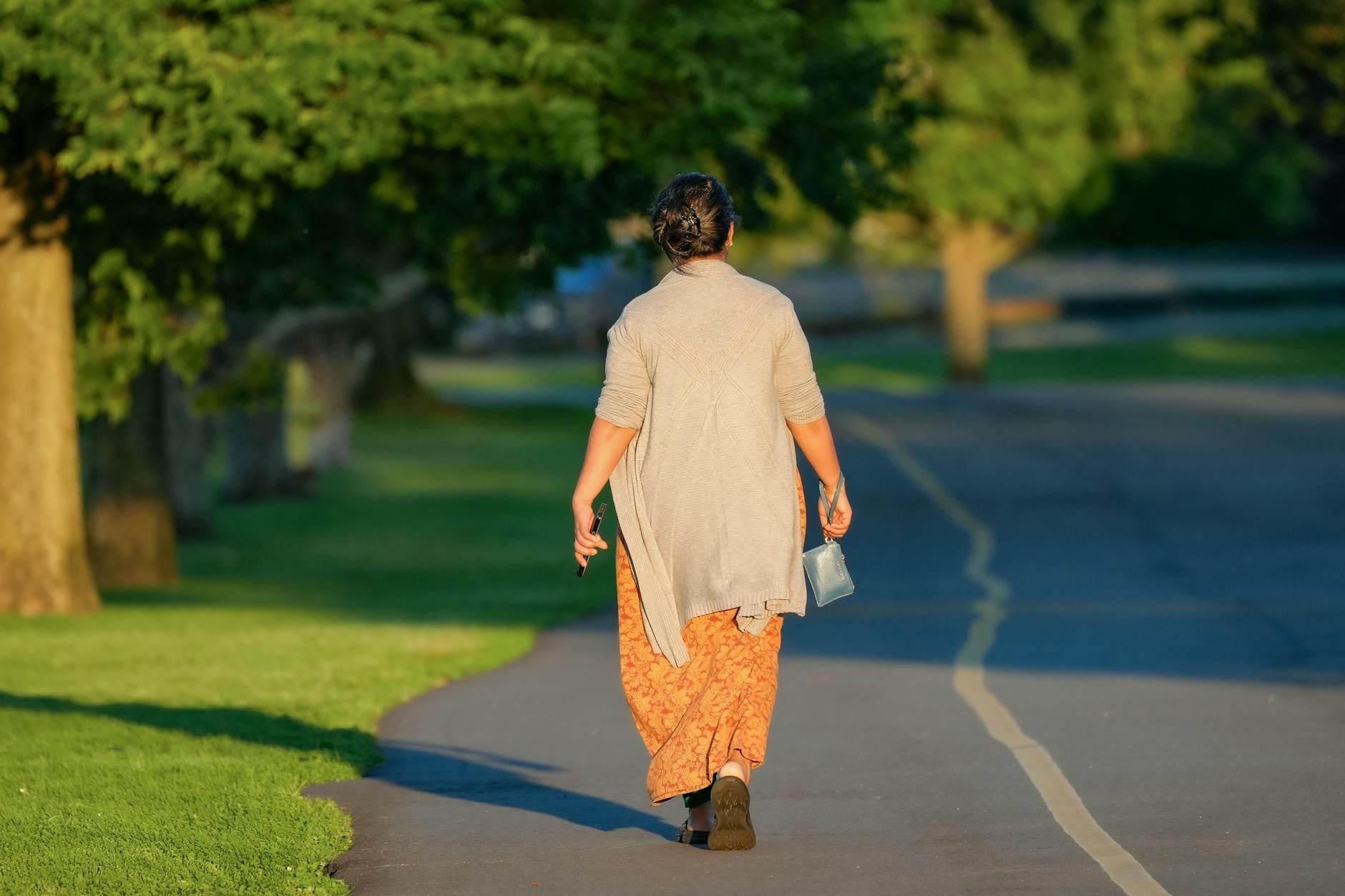 A woman walks down a tree-lined path in a park, enjoying a sunny morning.