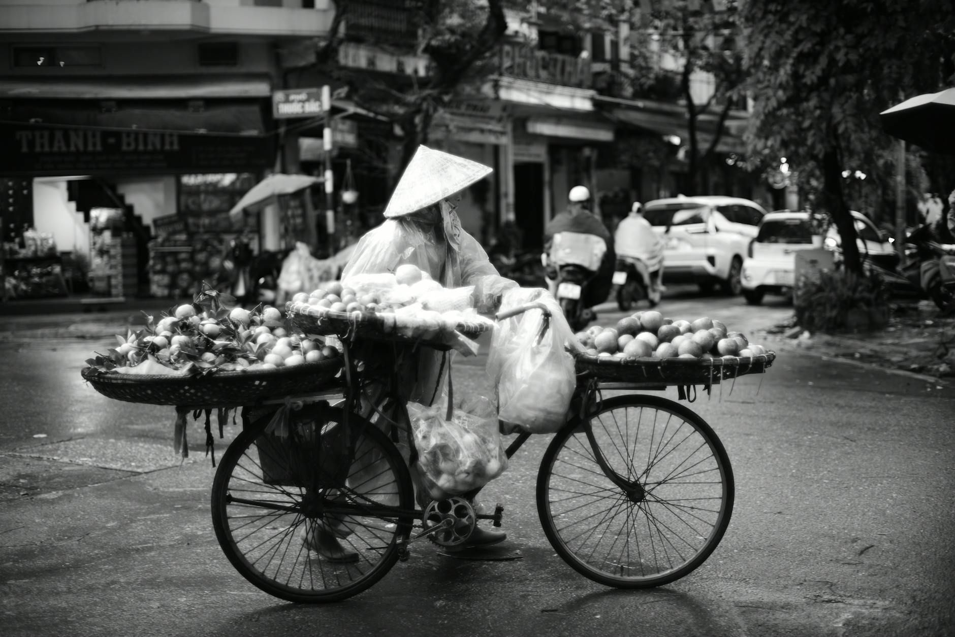 A street vendor with a traditional hat sells fruits from a bicycle in a bustling Vietnamese city.