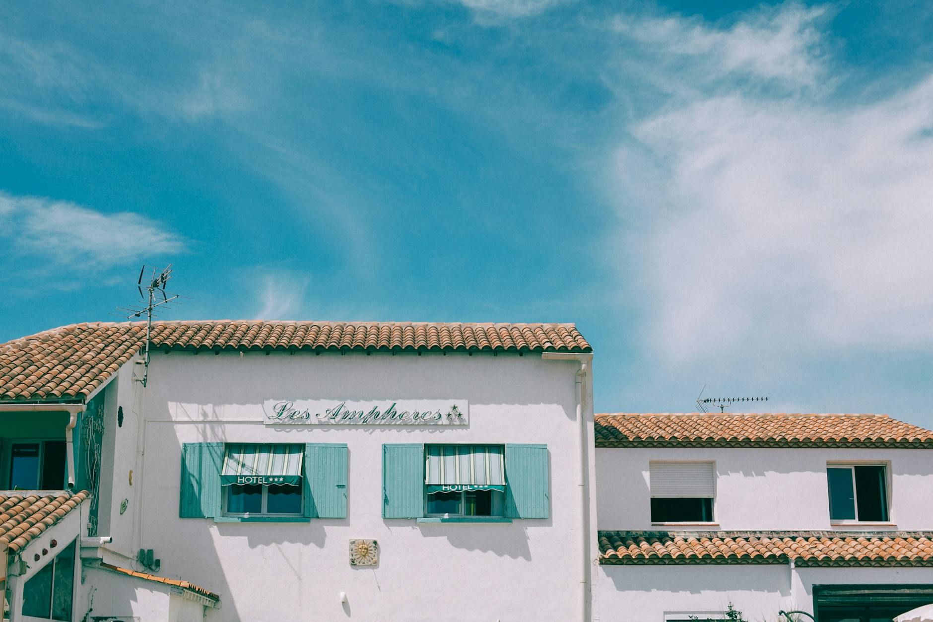 Exterior of cement building with wooden shutters on glass windows and red tiled roof under blue sky with clouds