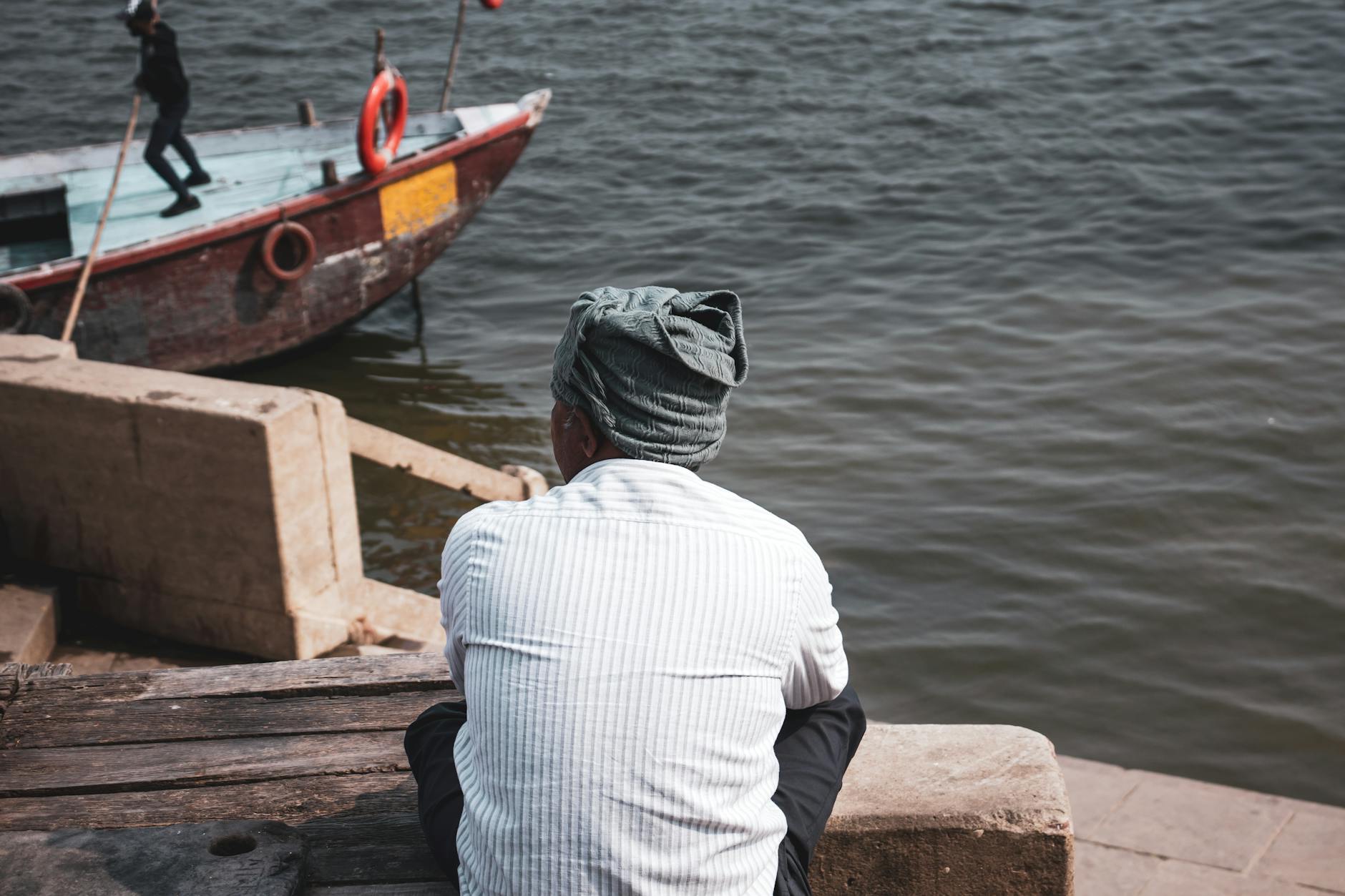 A man with headwear sits by the water, watching a boat under the sun.