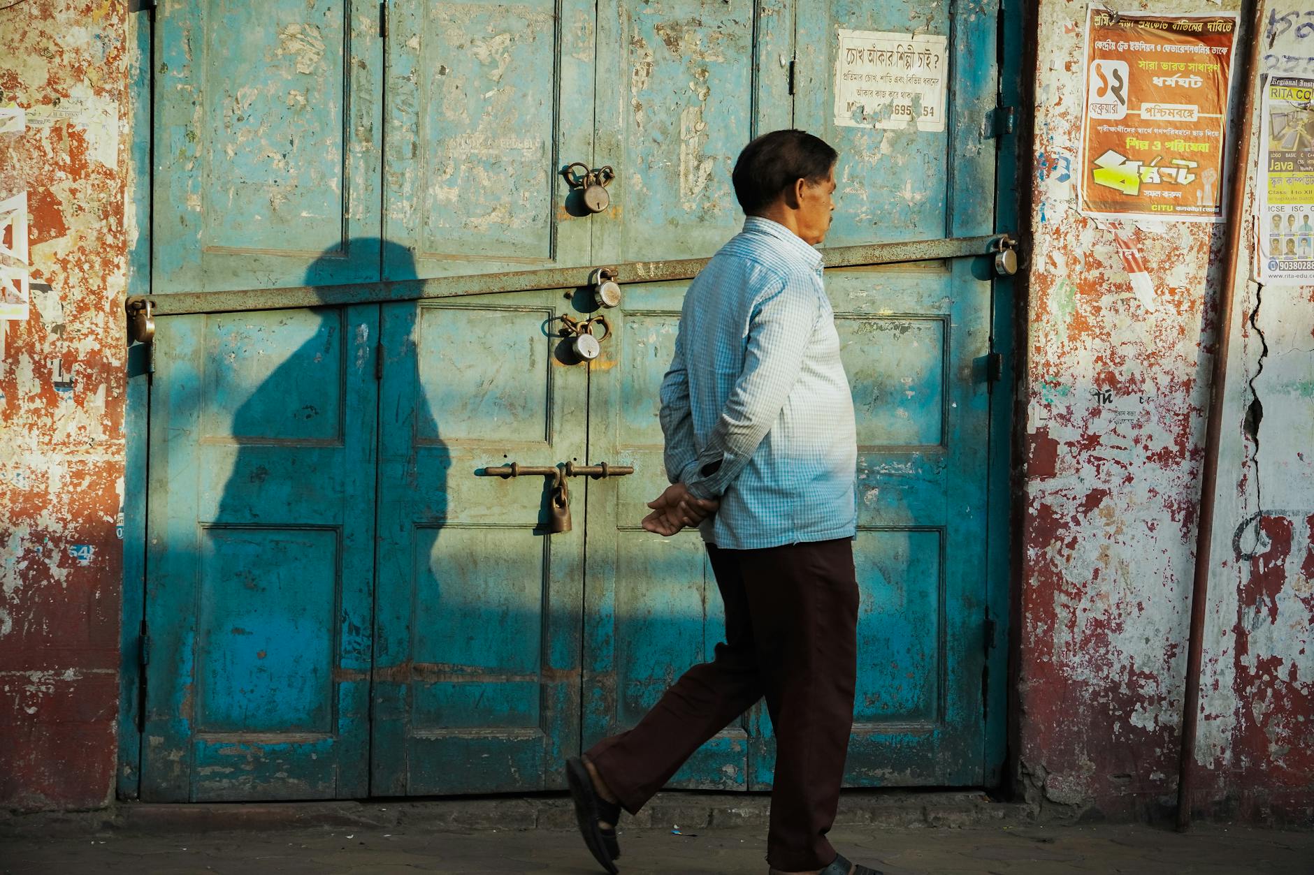 A man walking past worn, locked blue doors on a sunlit street in India.