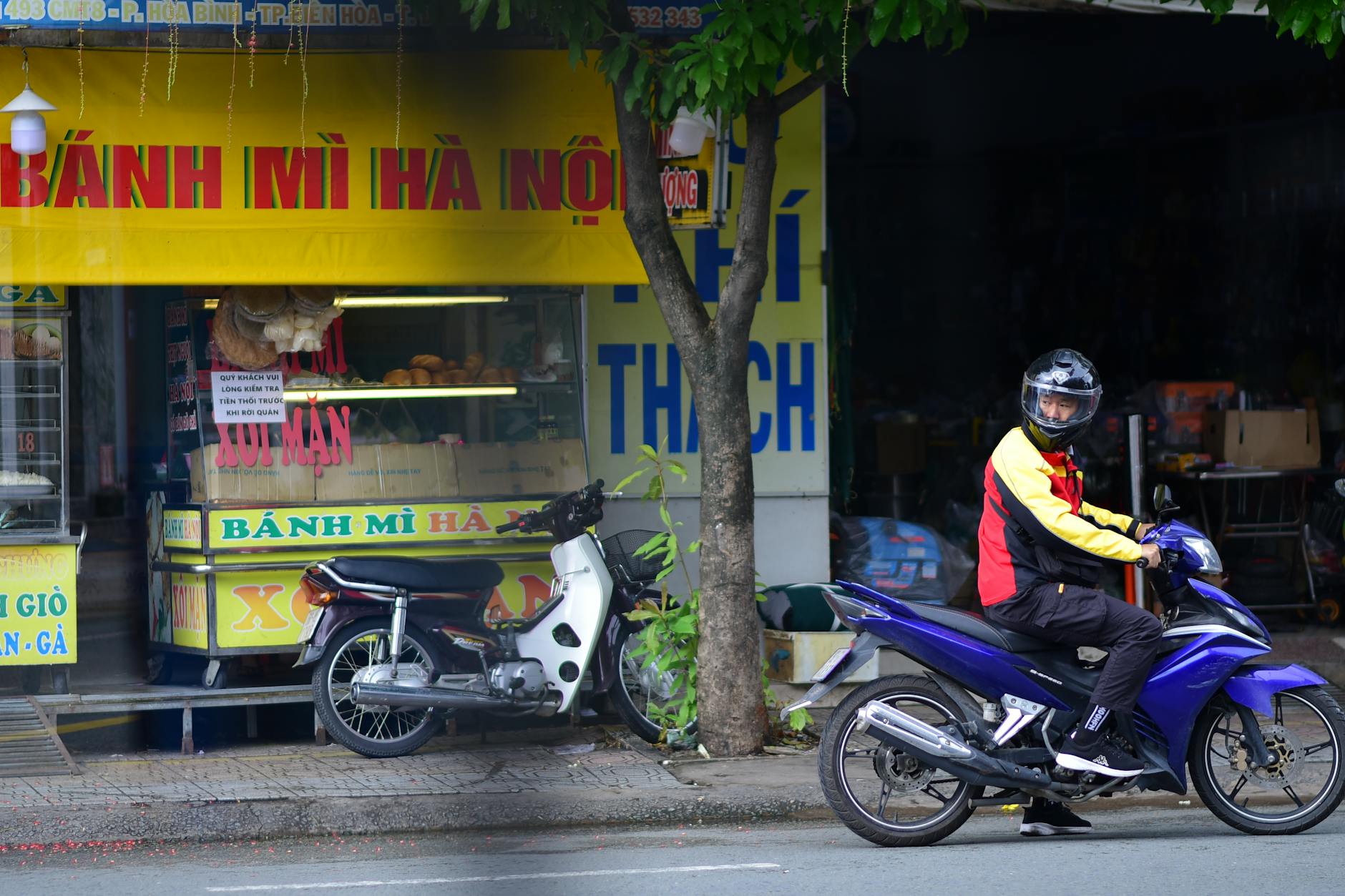 A motorcyclist pauses in front of a traditional Vietnamese Bánh Mì shop in an urban setting.