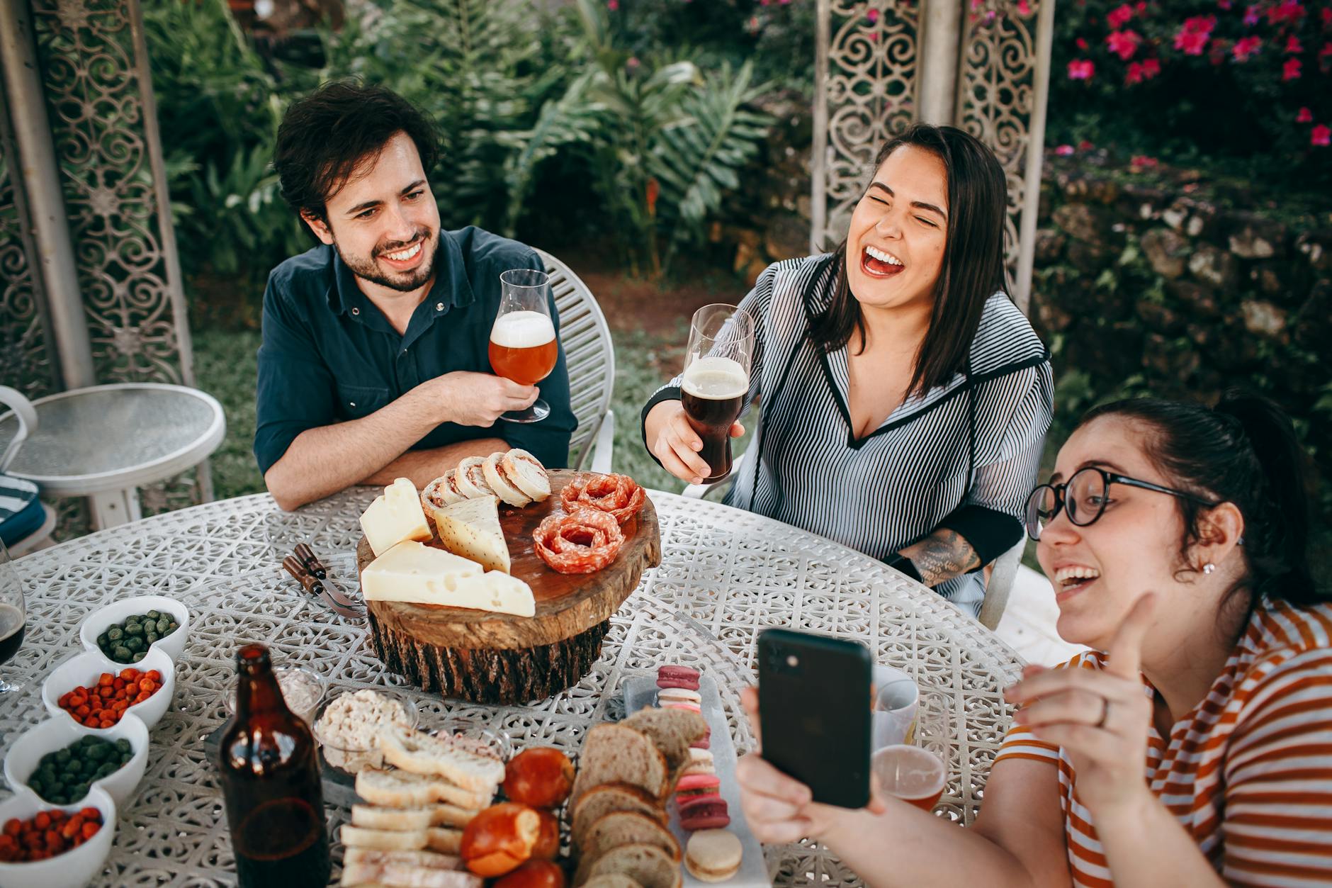A group of friends laughing and enjoying beer and snacks at an outdoor picnic table.