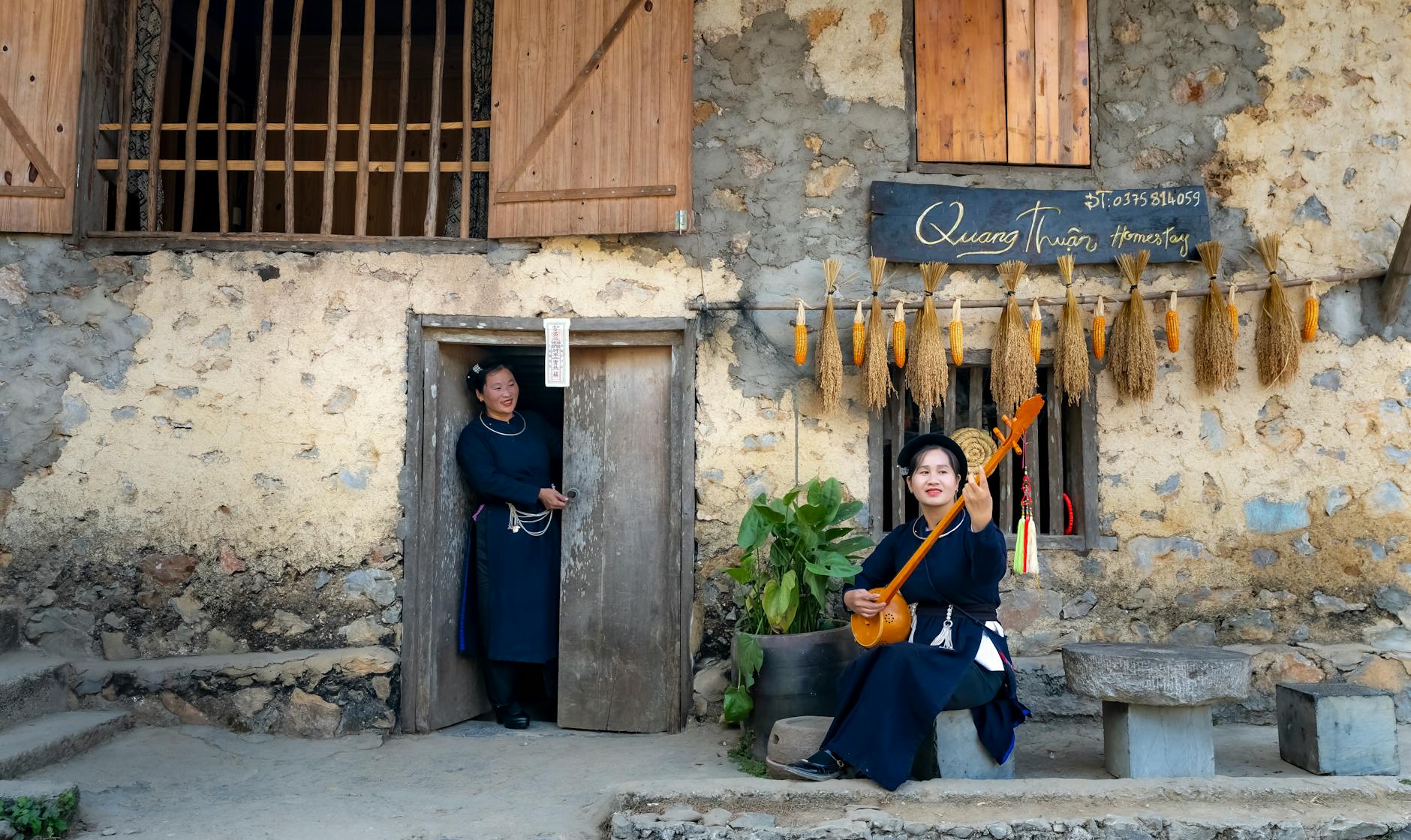 Two women in traditional attire play music at rustic Vietnamese homestay.