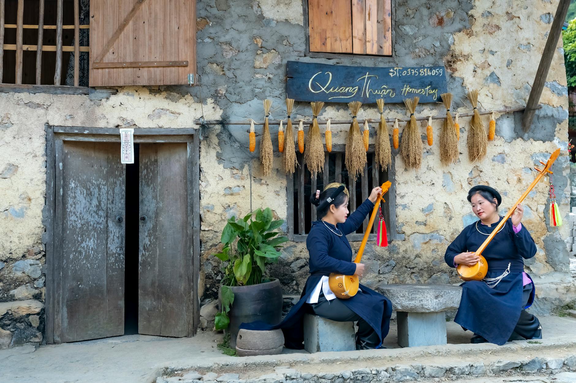 Two women in traditional attire playing string instruments outside a homestay.