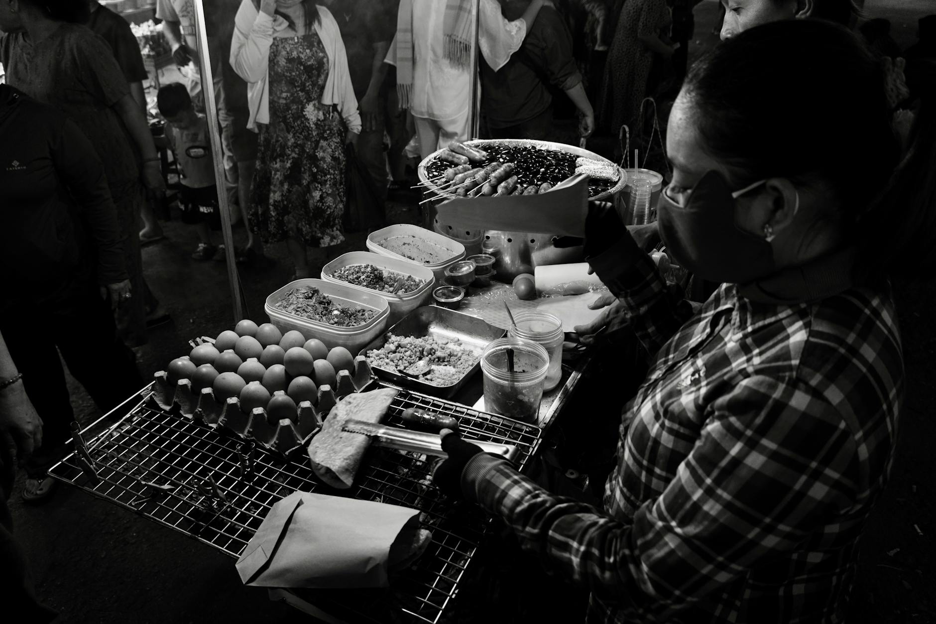 A woman serves street food at a bustling night market in monochrome.