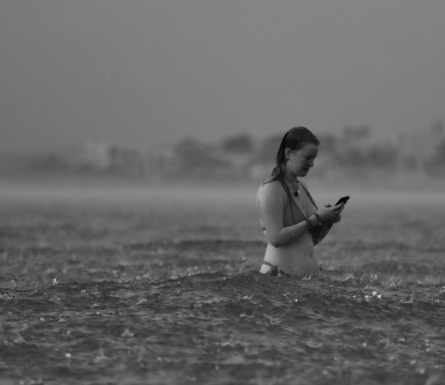 Black and white photo of a woman using her phone in the sea during a rainfall.
