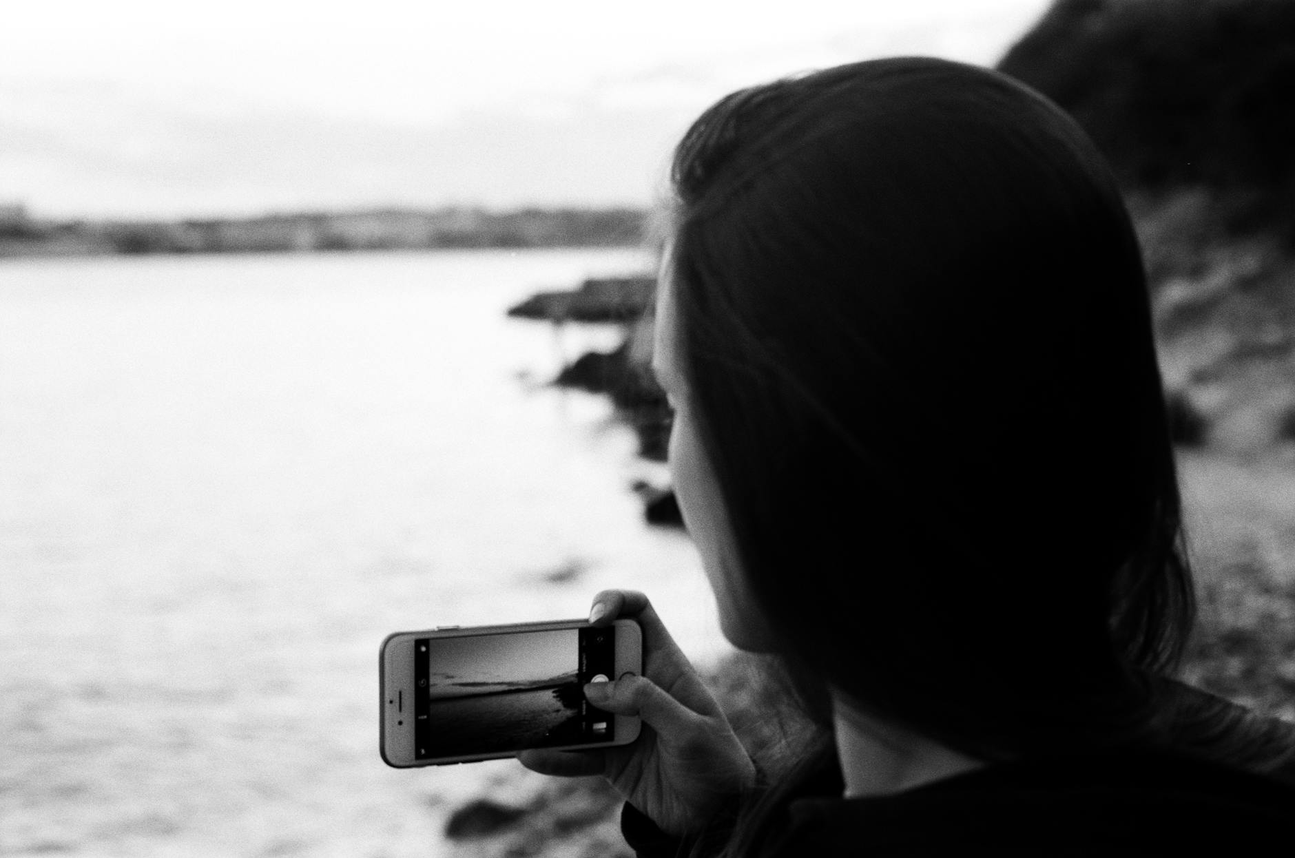 Black and white photo of a woman capturing the ocean view with her smartphone on a serene day.