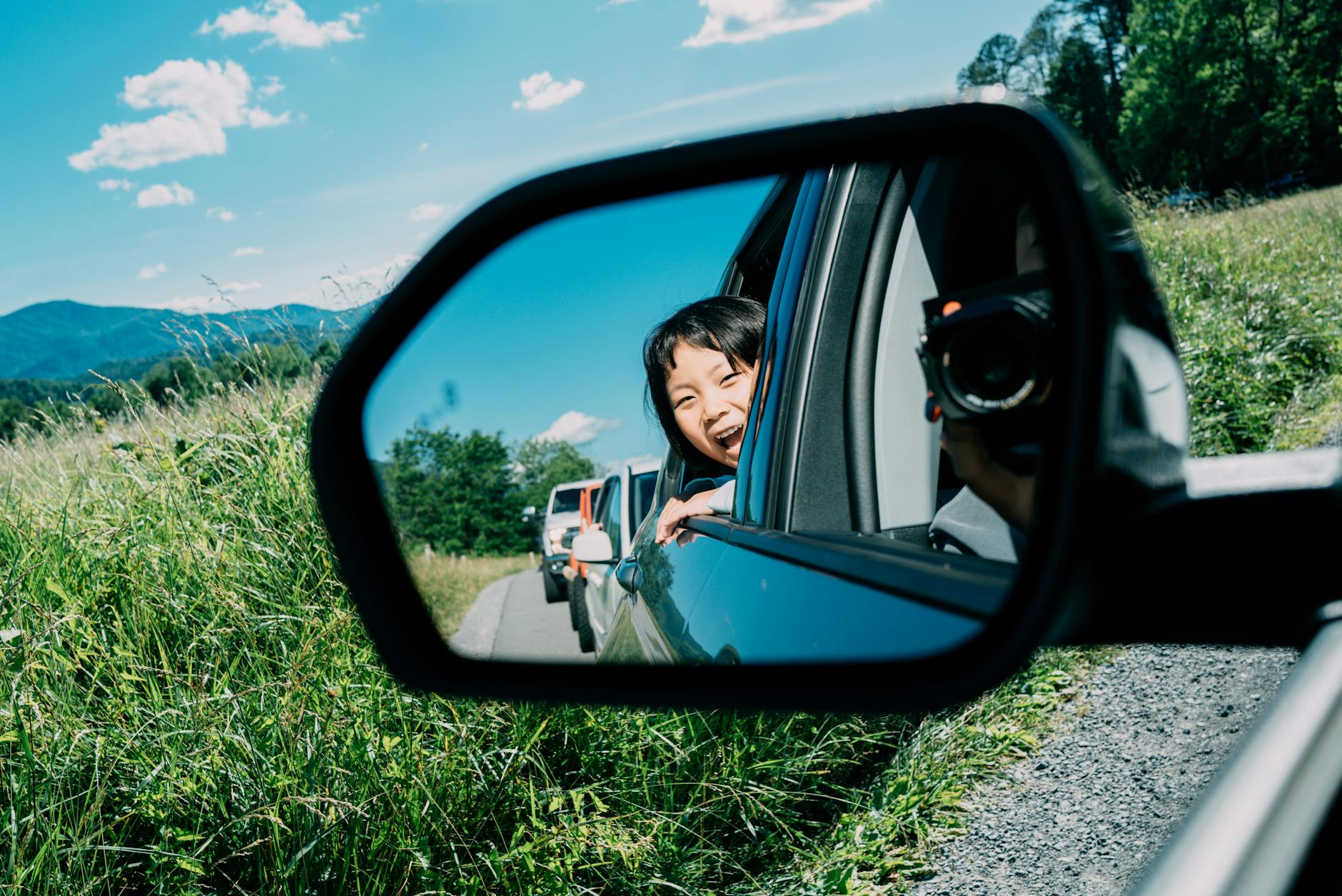 A smiling child looks out of a car's side-view mirror during a sunny road trip through lush greenery.
