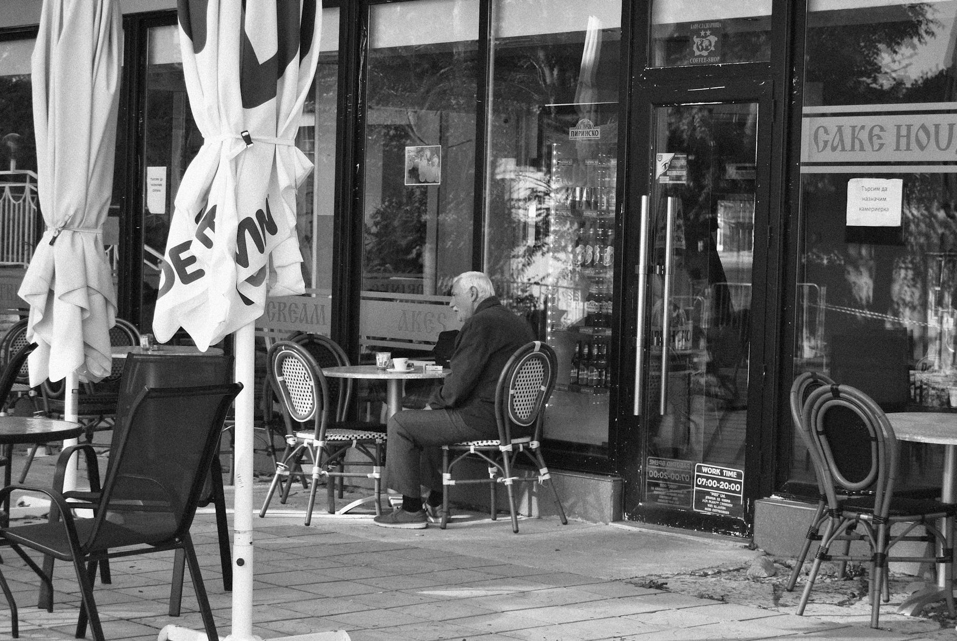 Black and white photo of an elderly man sitting alone at an outdoor café table.
