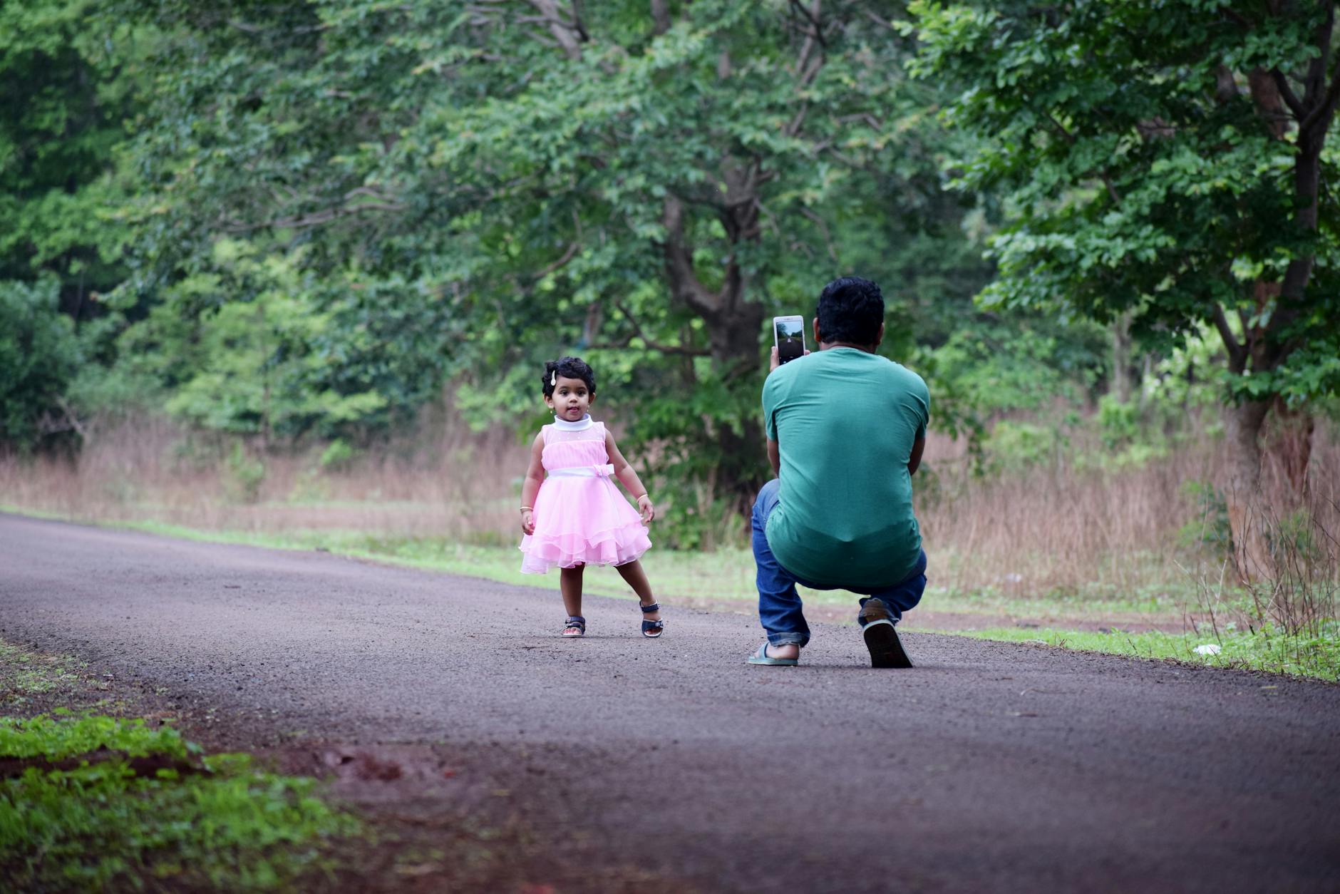 A father takes a photograph of his daughter in a pink dress on a scenic outdoor path.