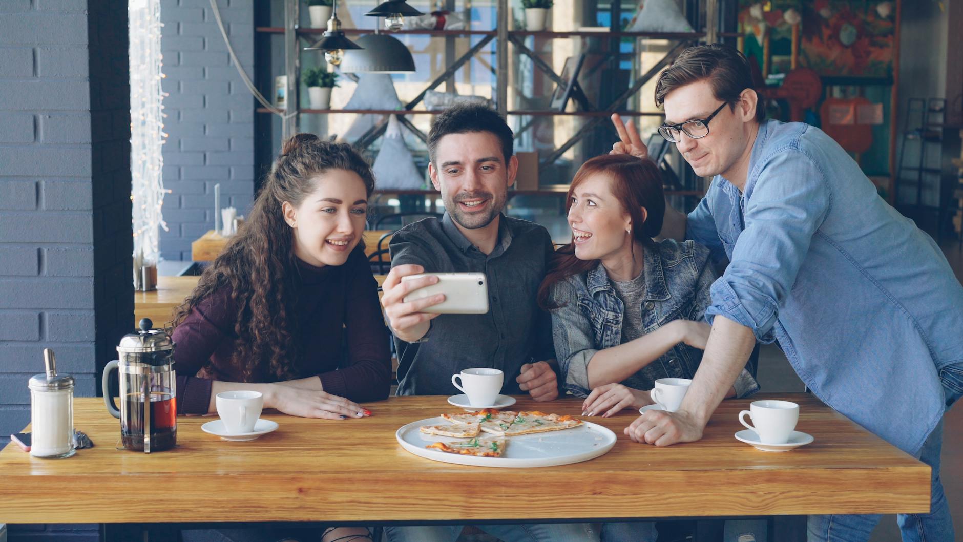 Four friends enjoying pizza and coffee while taking a selfie in a cozy cafe.