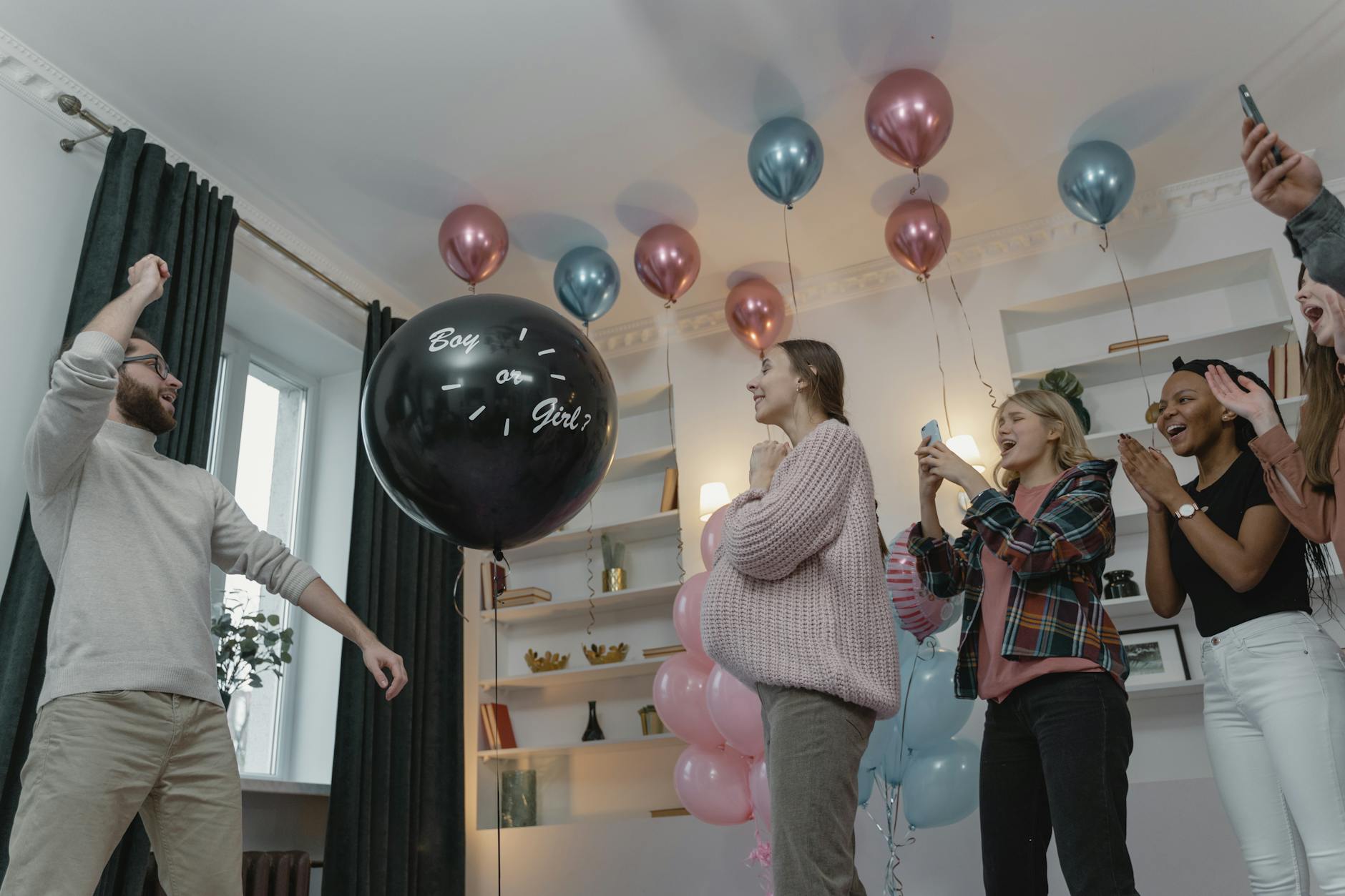 Group of friends celebrating a gender reveal party with balloons and excitement indoors.