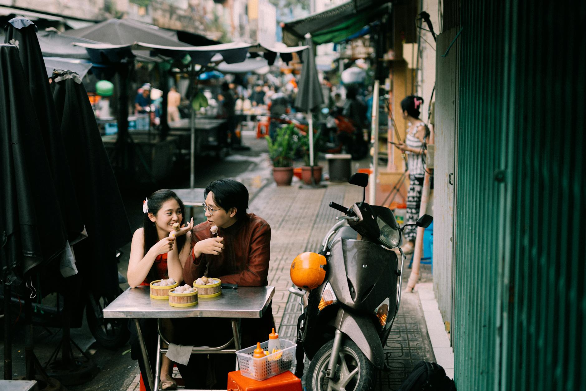 A couple enjoys a casual meal at an outdoor market, surrounded by street vendors and lively atmosphere.