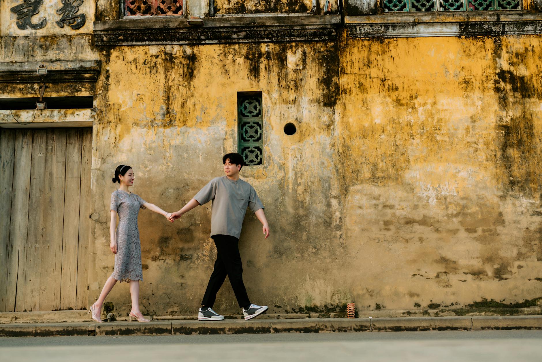 A happy couple holding hands while walking along a rustic wall on a sunny day.