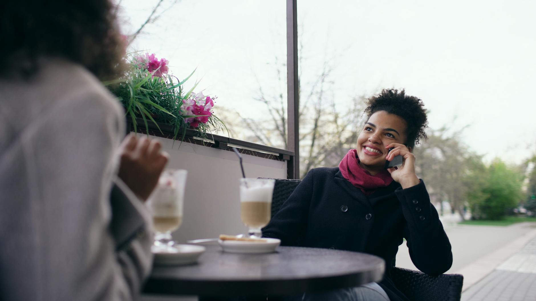 A joyful woman chats on the phone while enjoying coffee outdoors.
