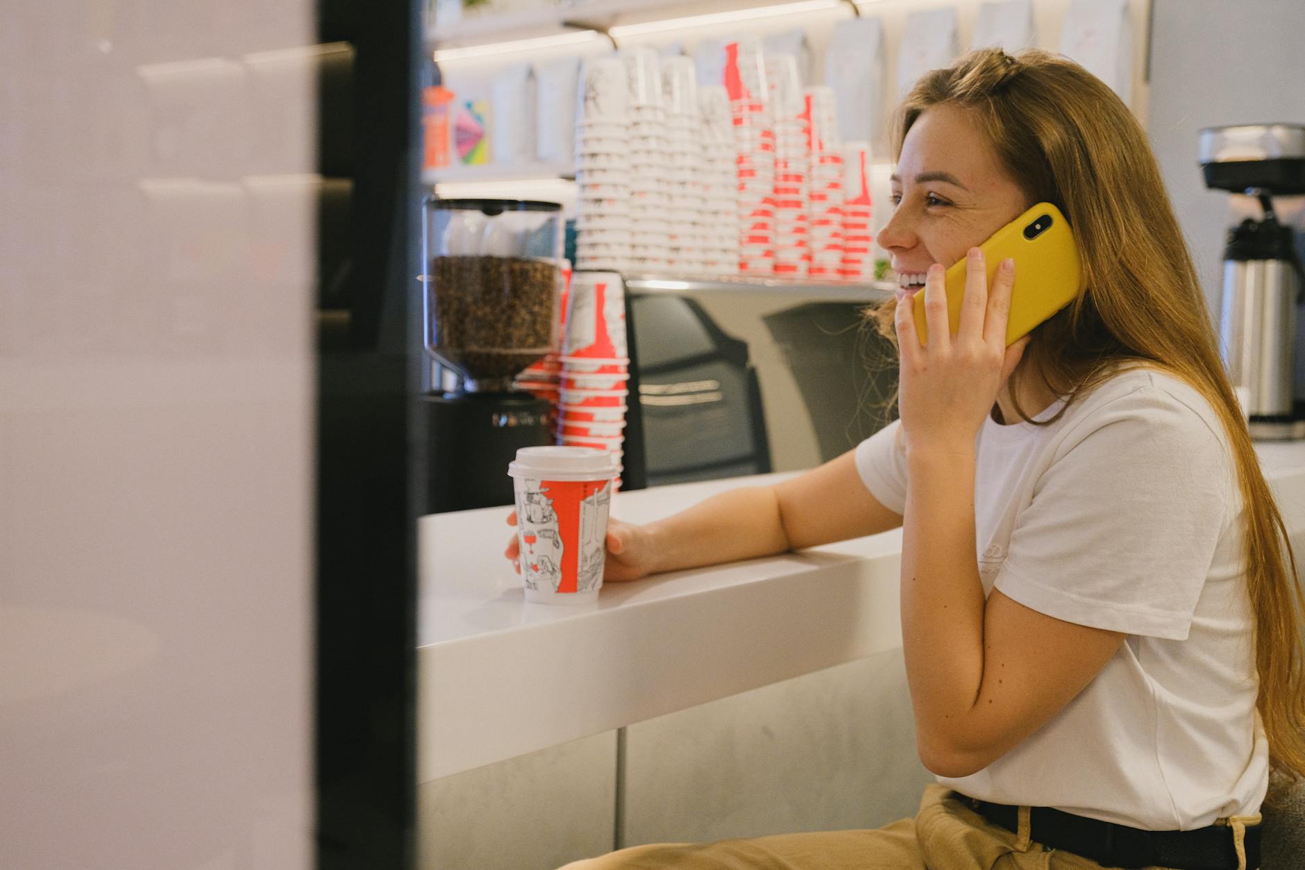 Smiling woman in coffee shop talking on smartphone, enjoying a hot drink.