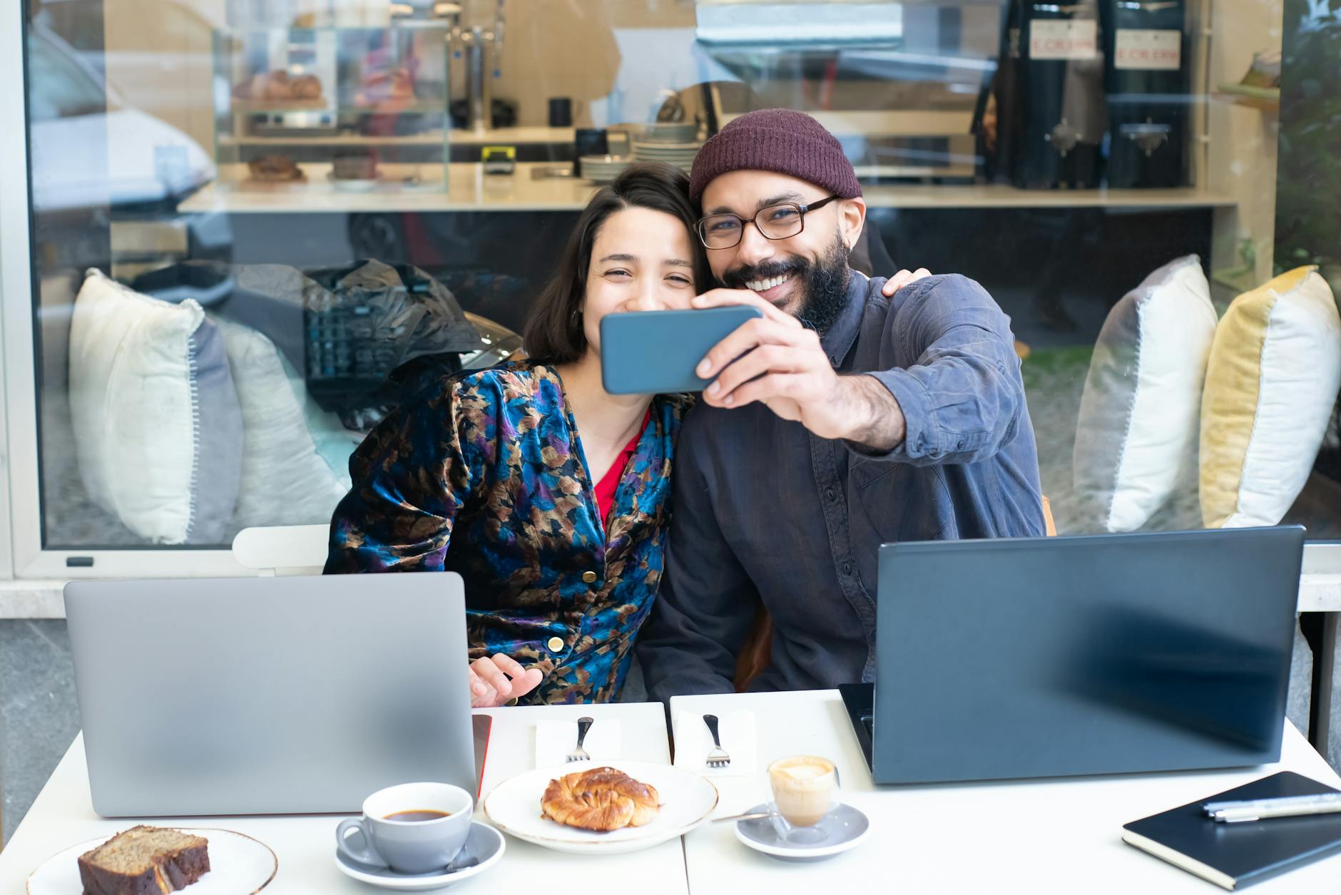 A smiling couple taking a selfie in a café while working on laptops, enjoying coffee and pastries.