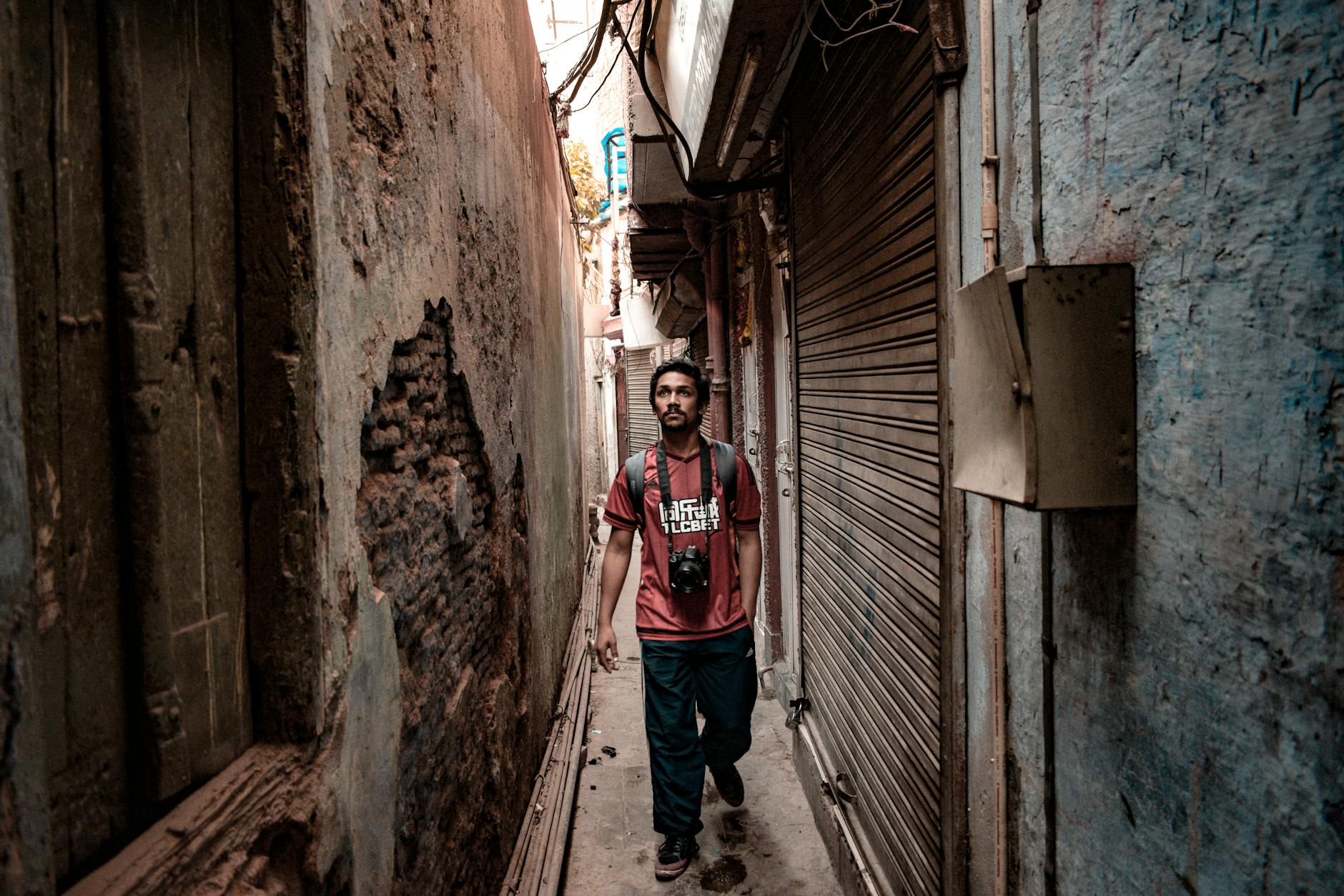A young man with a camera walks through a narrow alley in an Indian urban setting, capturing street life.