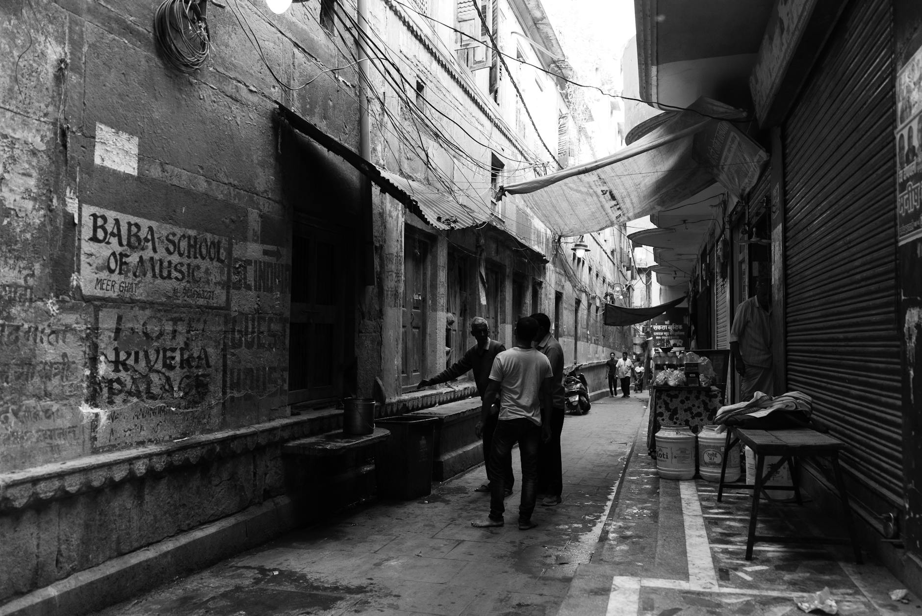 A black and white photograph of a narrow alleyway bustling with life in an Indian city.