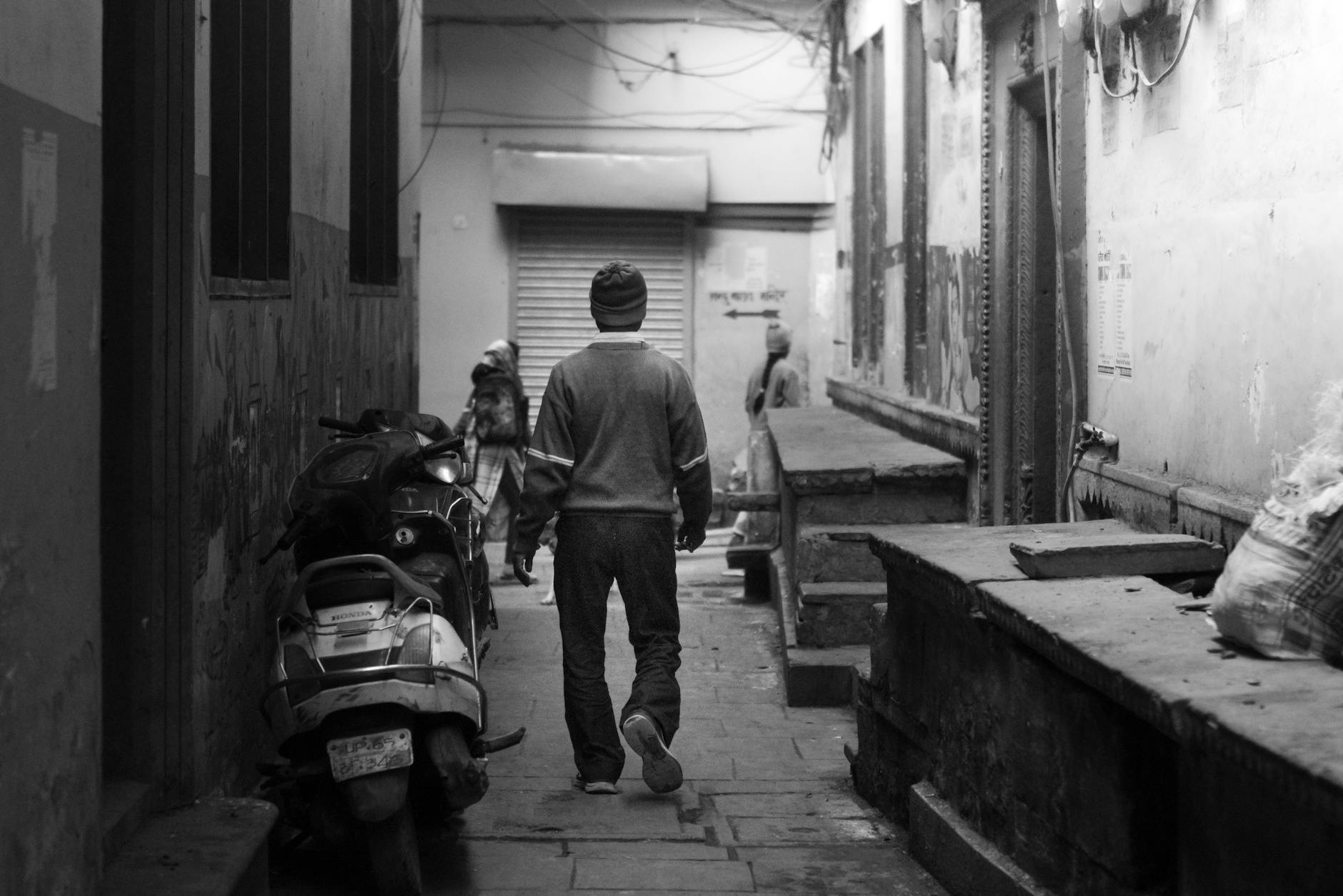 A black and white image of a man walking down a narrow street in Varanasi, India.