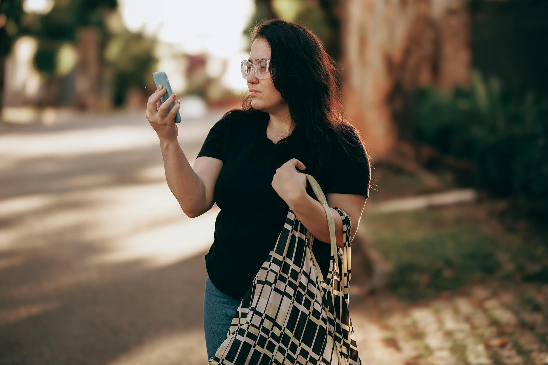 A woman stands on a sunny sidewalk, using her phone, carrying a patterned bag.