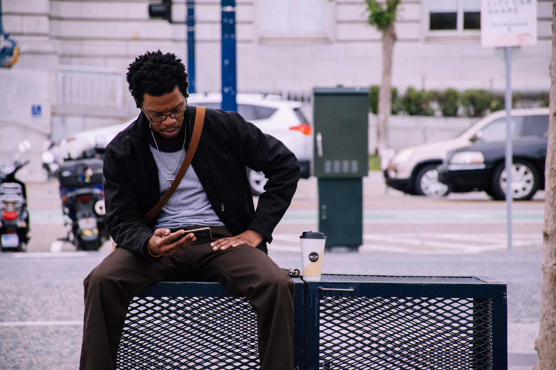 Man sitting on a street bench in San Francisco, checking his phone with a coffee cup nearby.