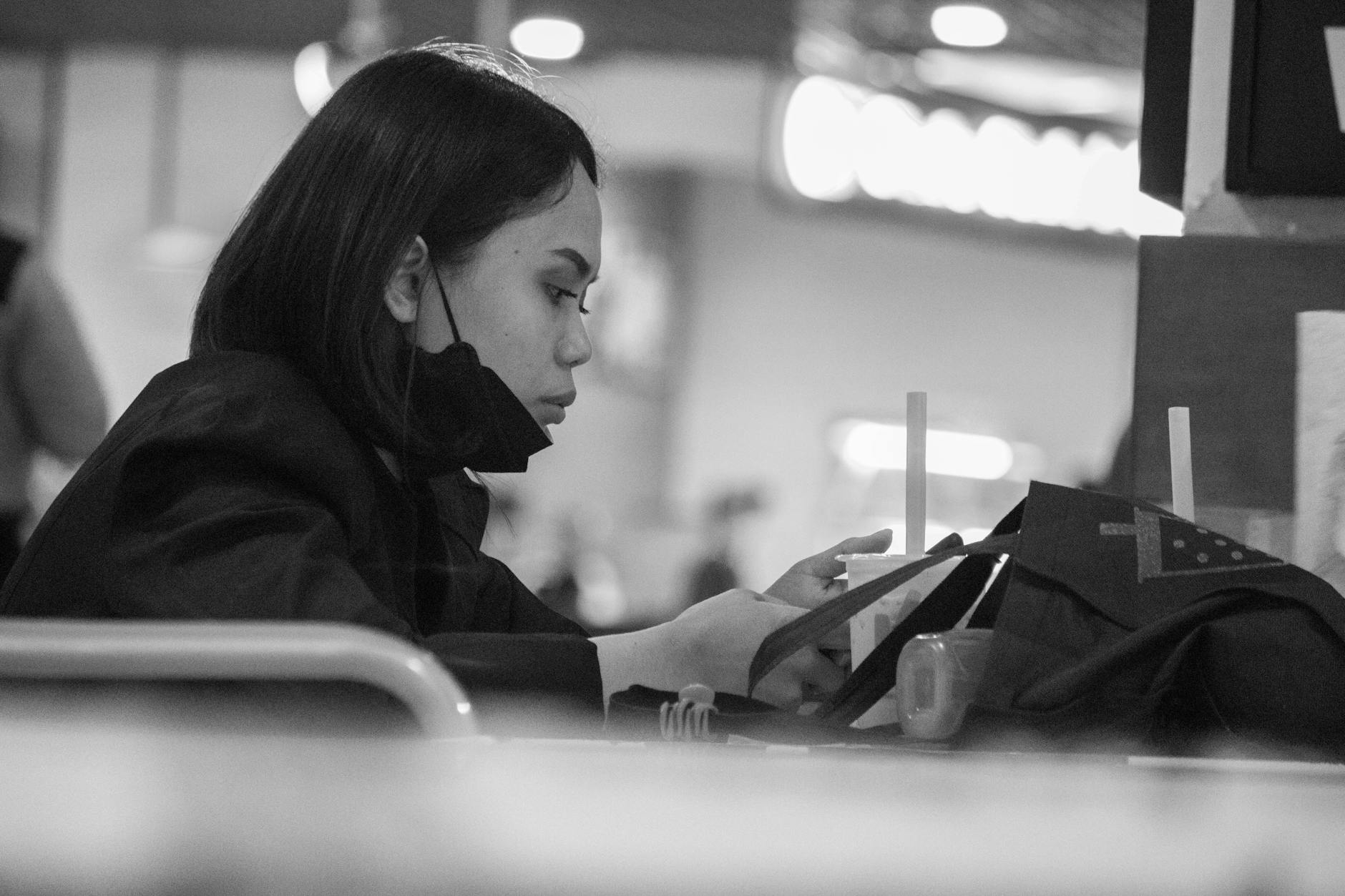 Black and white candid shot of a young woman using her phone at a food court, seated at a table.