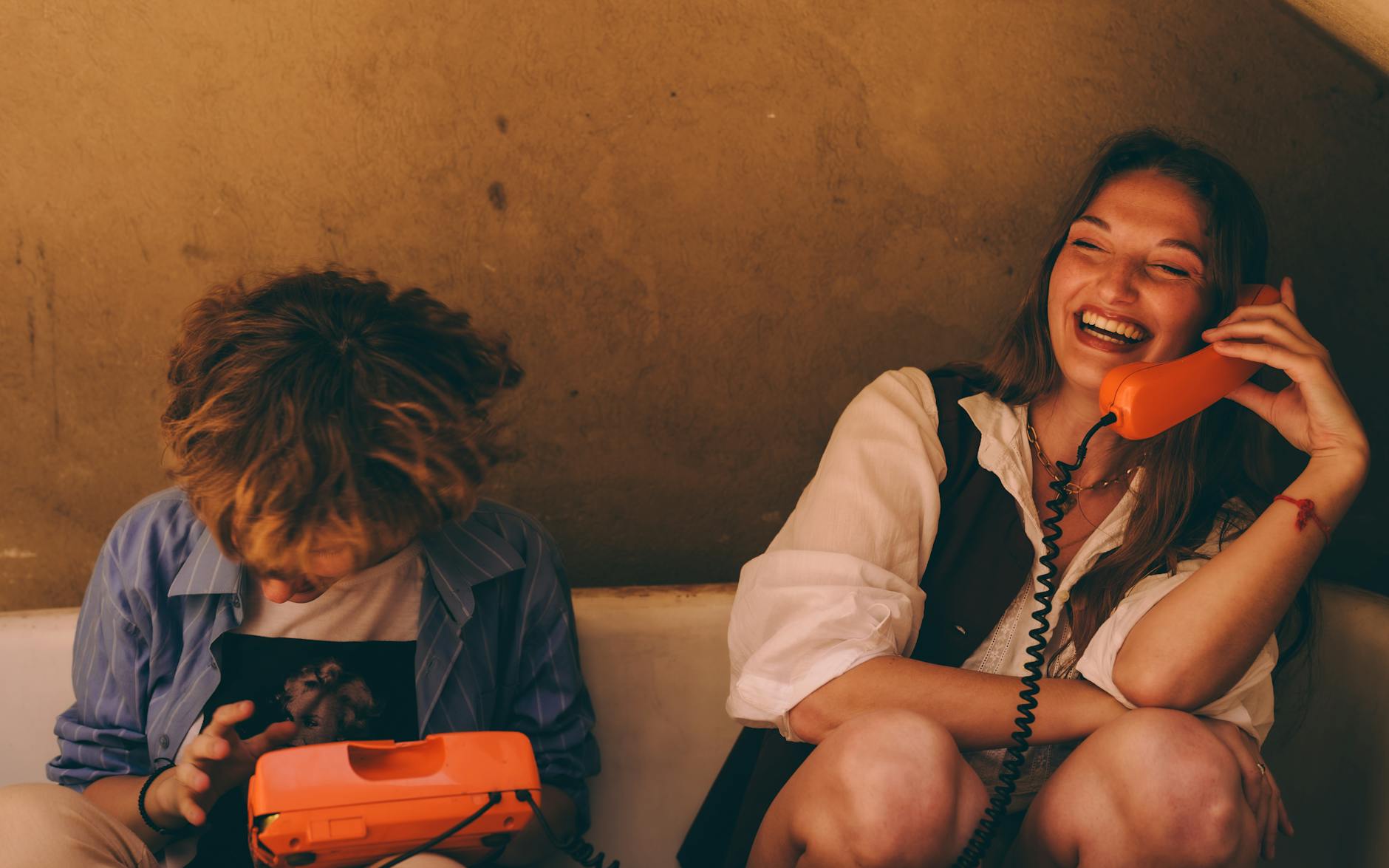 Two young adults enjoying a lively conversation on vintage orange corded phones indoors.