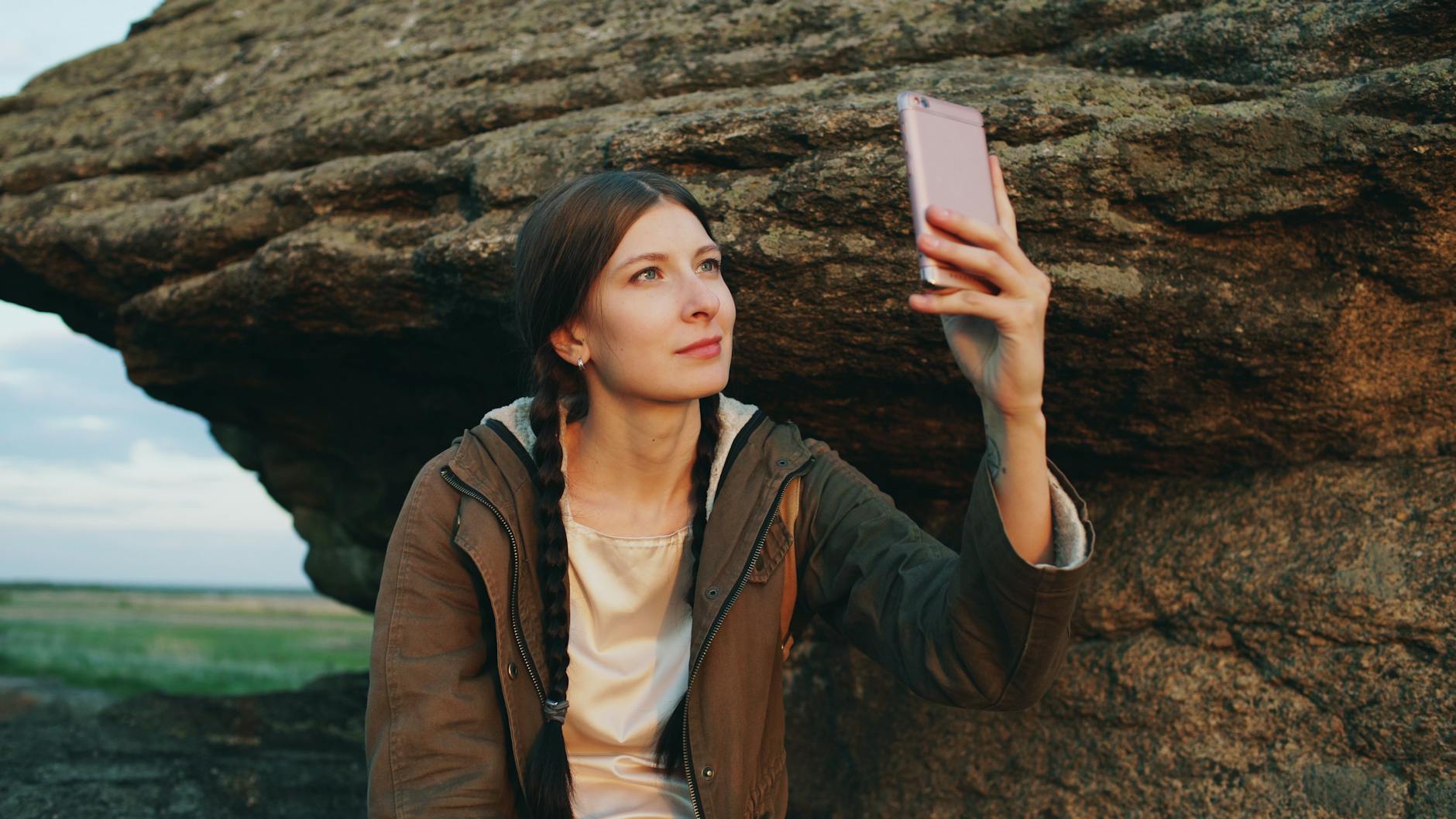 A woman taking a selfie on her smartphone near a large rock formation outdoors.