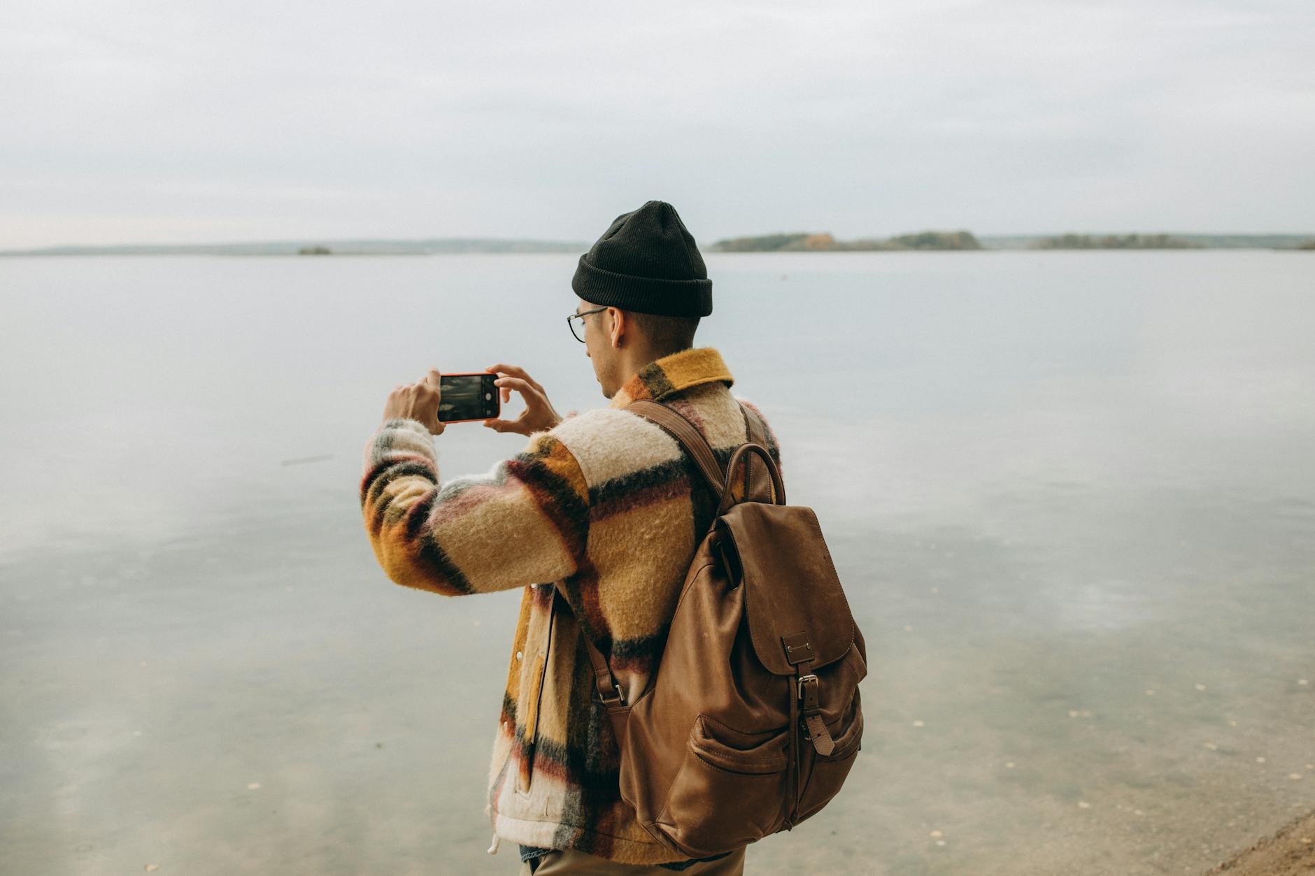 Back view of a man capturing a lake with a smartphone, in a peaceful outdoor setting.