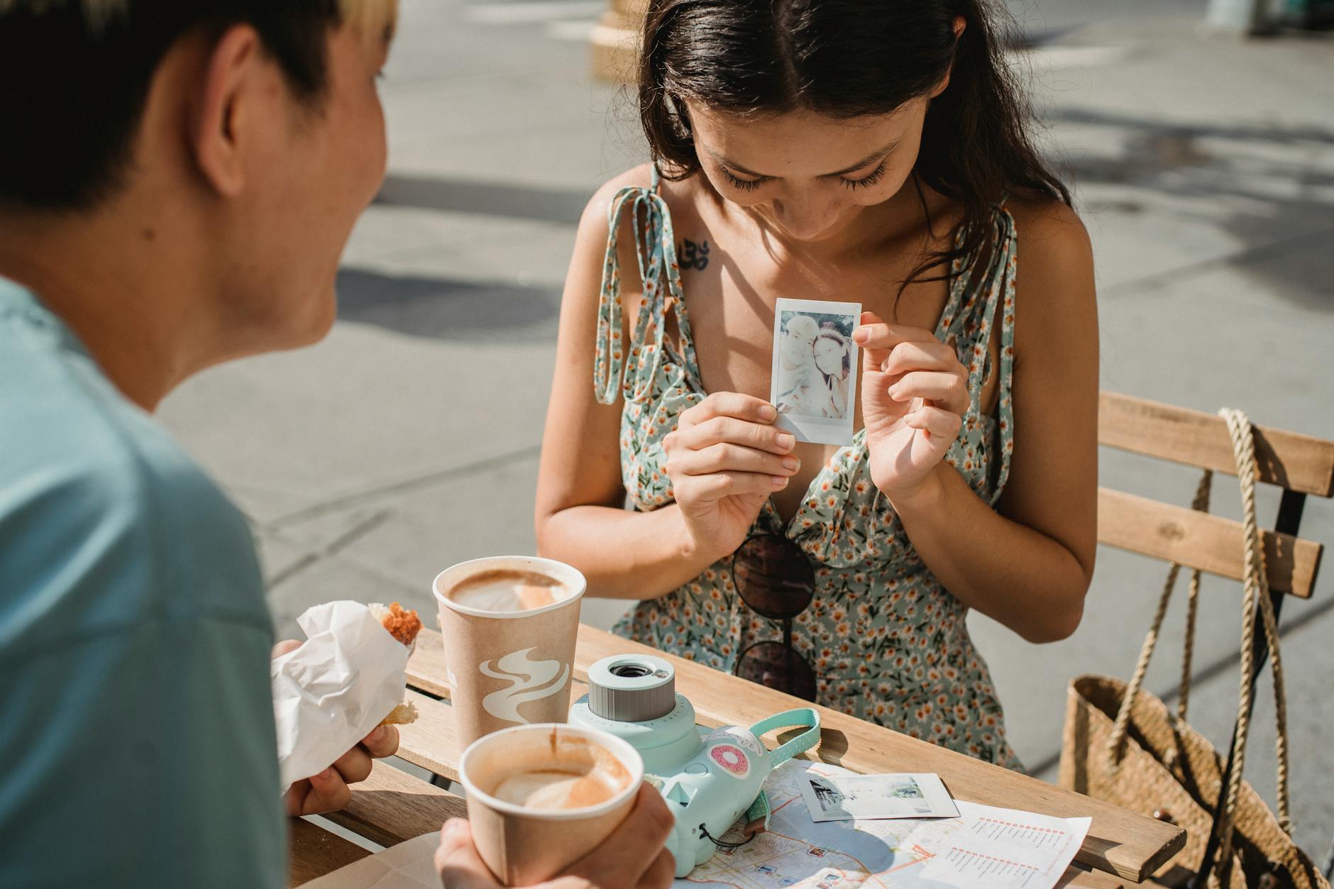 Crop smiling ethnic female showing instant photo to anonymous partner with disposable glass of cappuccino at cafe table in sunlight