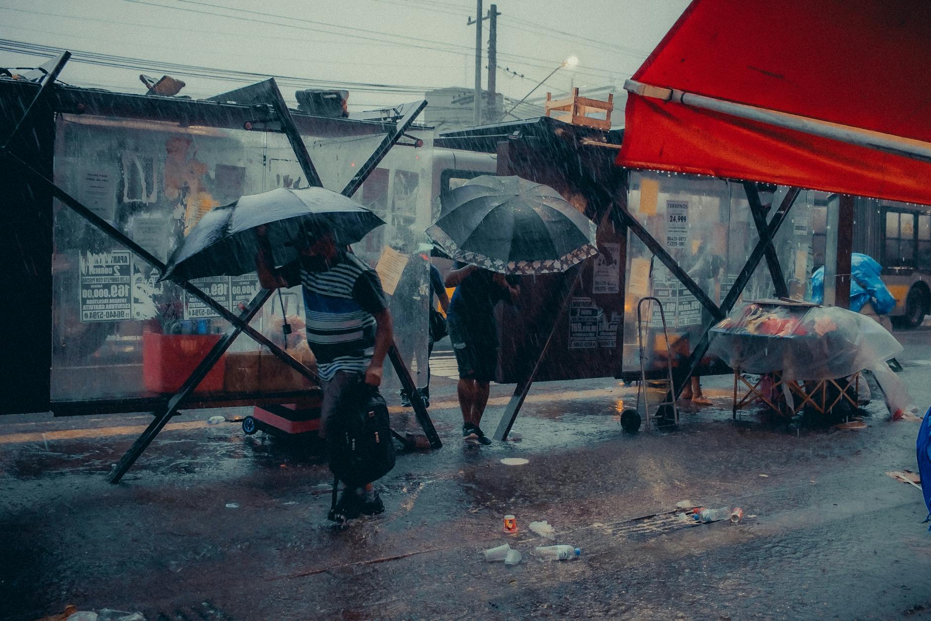 People with umbrellas at a city bus stop during heavy rain.