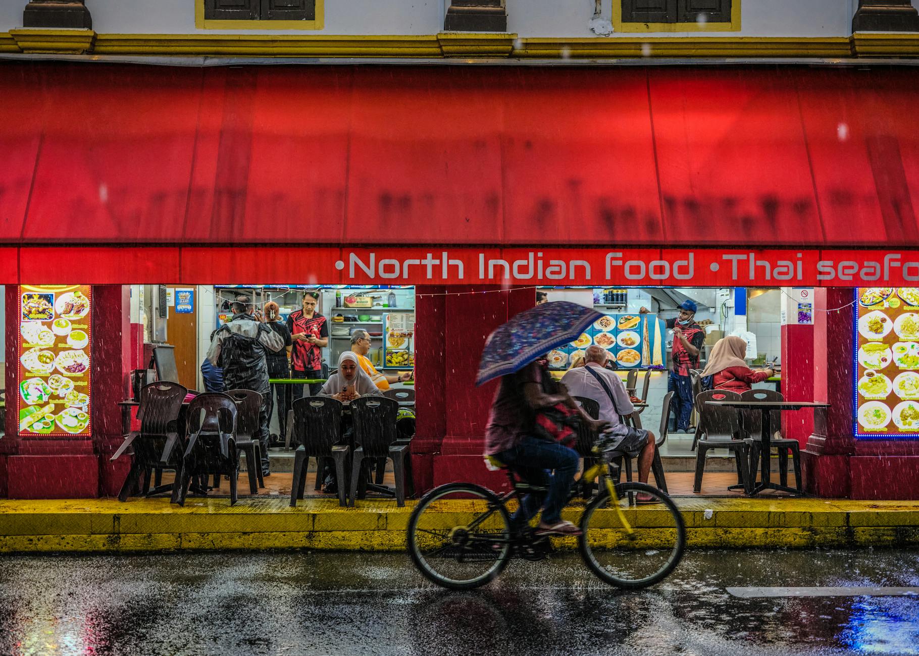 Street view of a North Indian restaurant with a cyclist in rain, capturing vibrant night life.