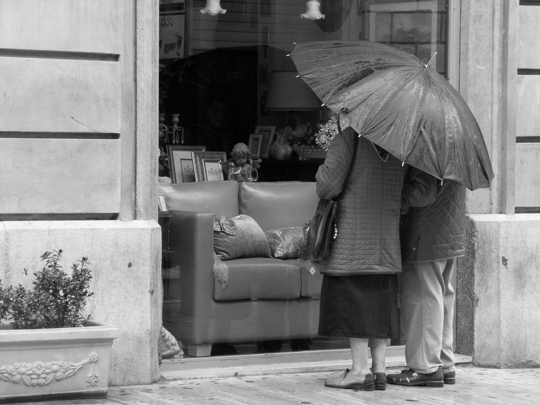 A couple stands under an umbrella, gazing into a store window on a rainy day.