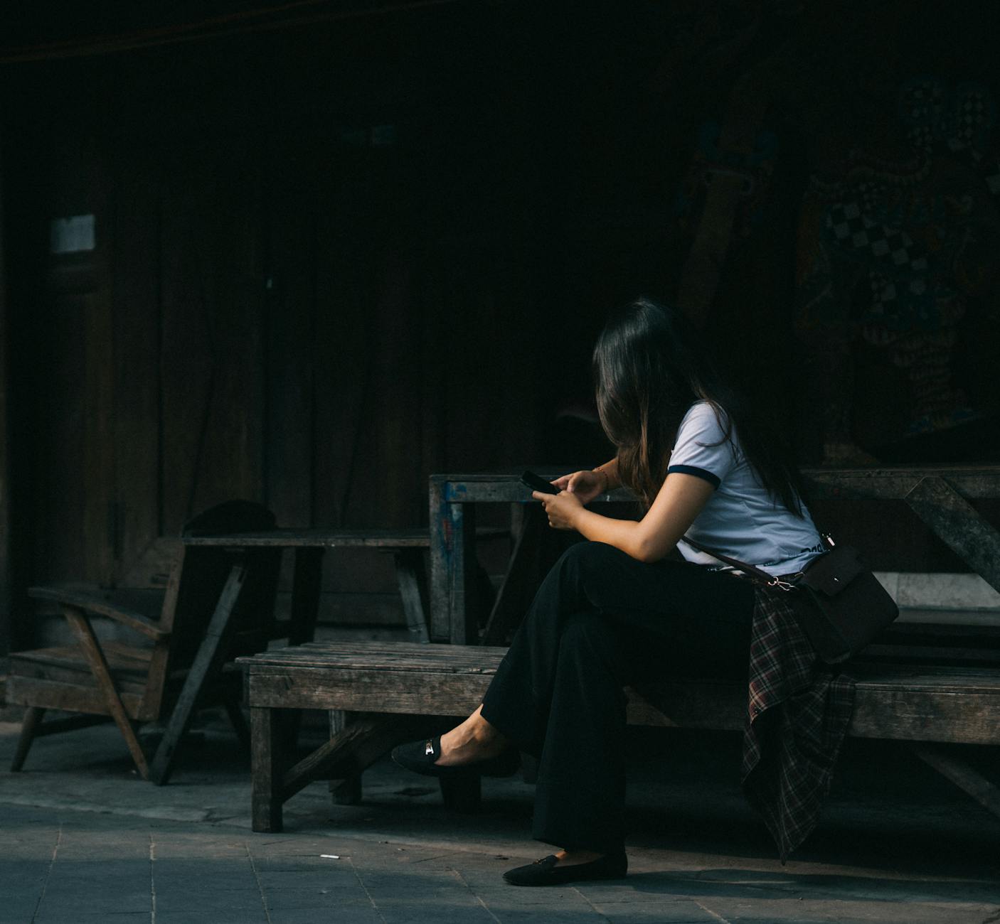 A young woman in casual attire is focused on her phone while sitting on a rustic wooden bench.