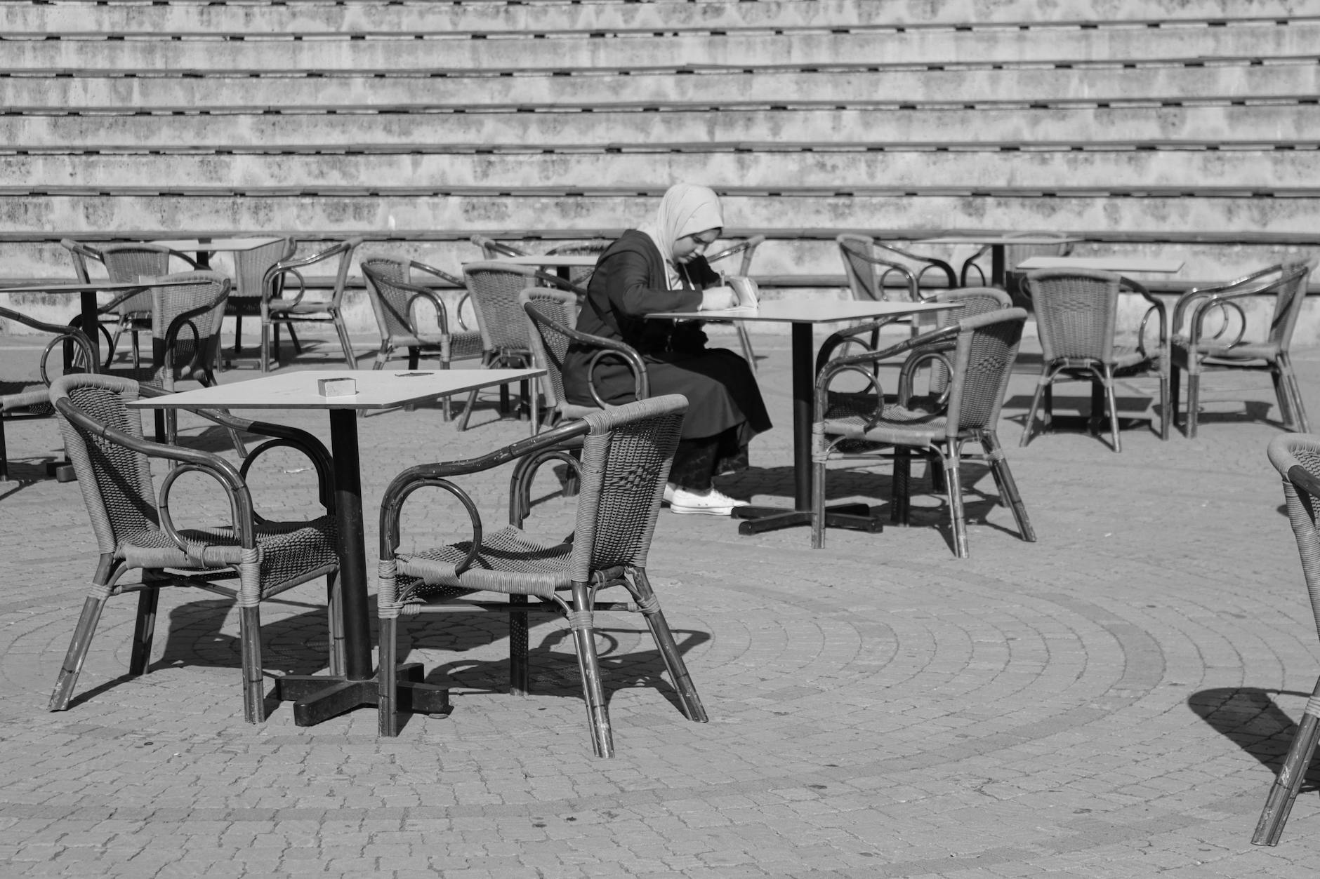 Black and white photo of a woman in hijab sitting at a table outdoors, capturing solitude.