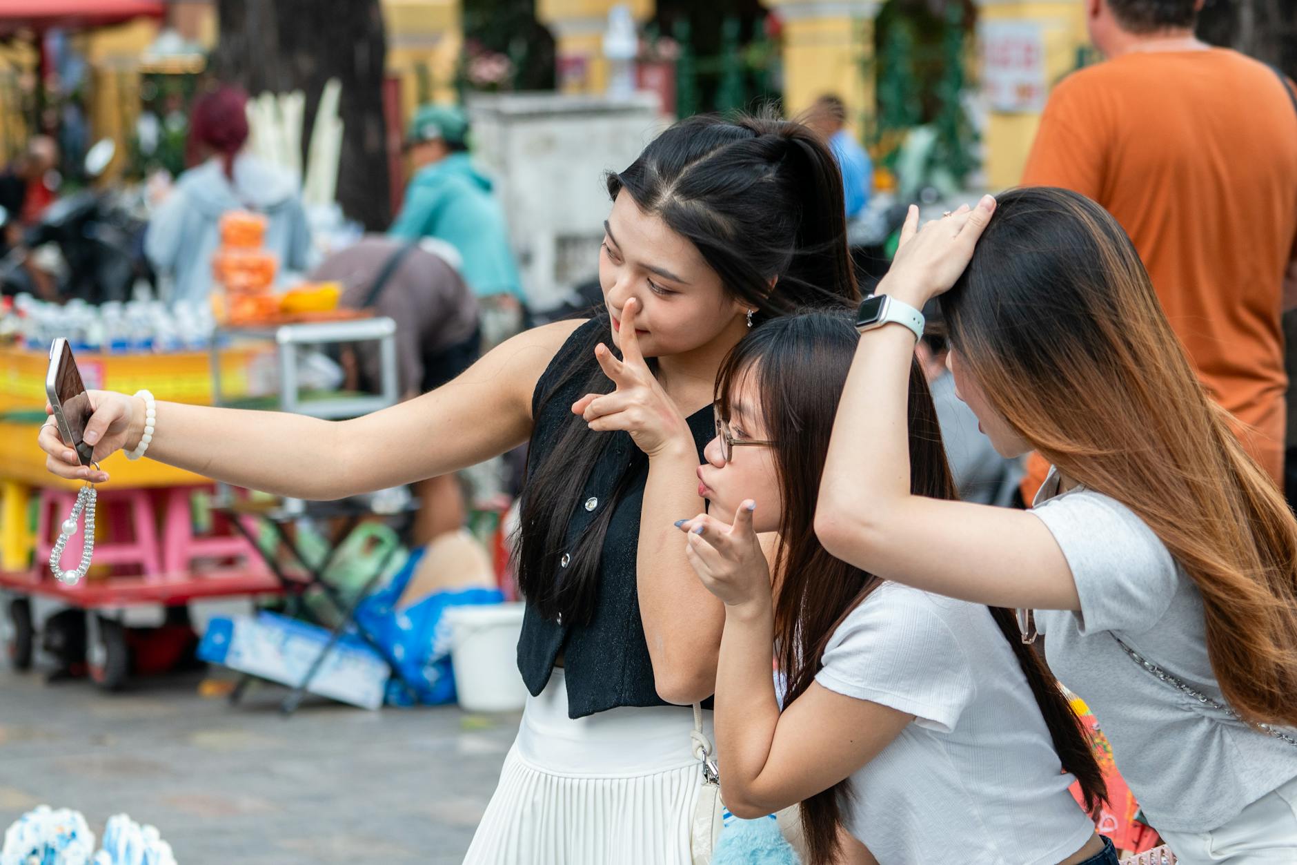 Three young women take a selfie together while enjoying a vibrant outdoor market scene.