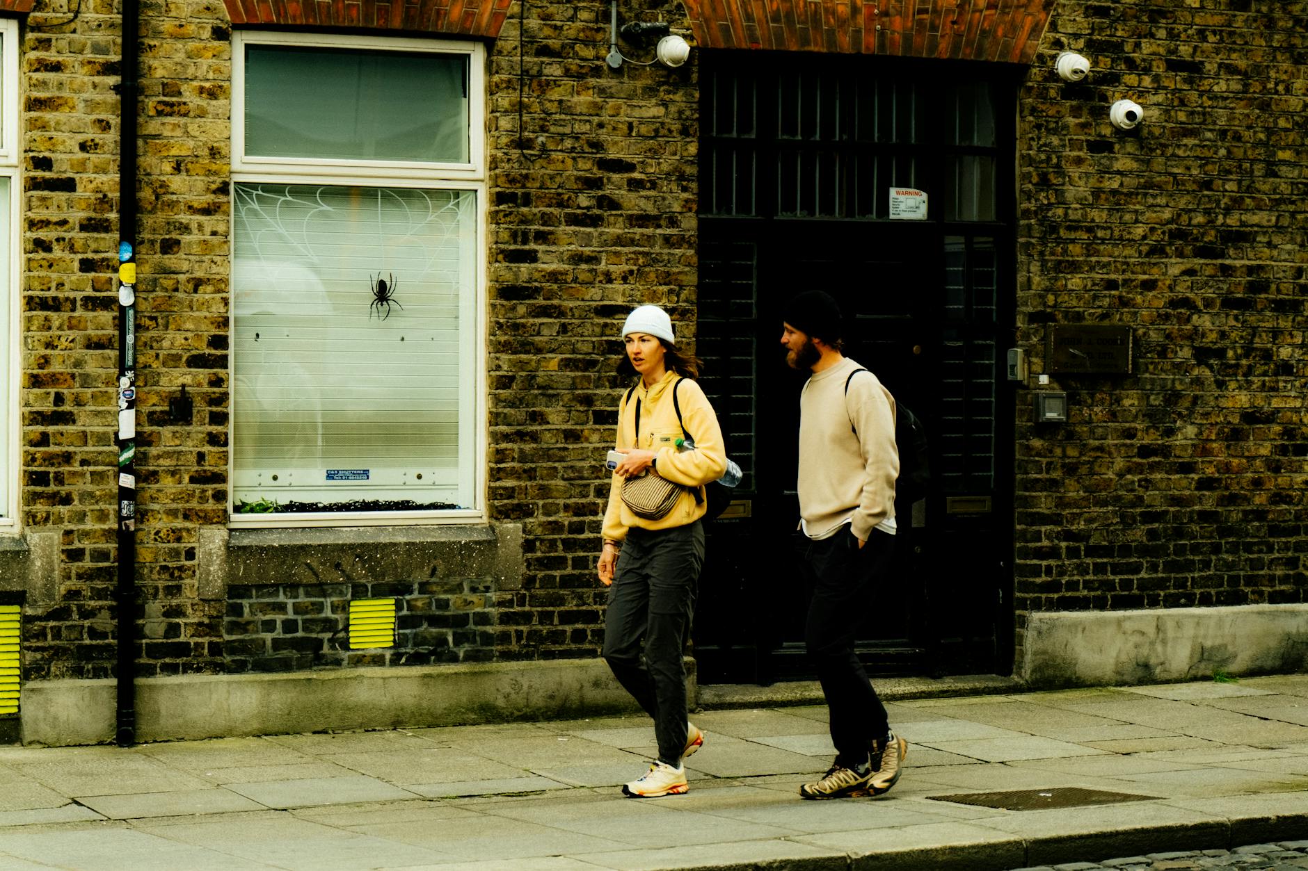 Two adults walking past a brick building in an urban setting, captured during the day.