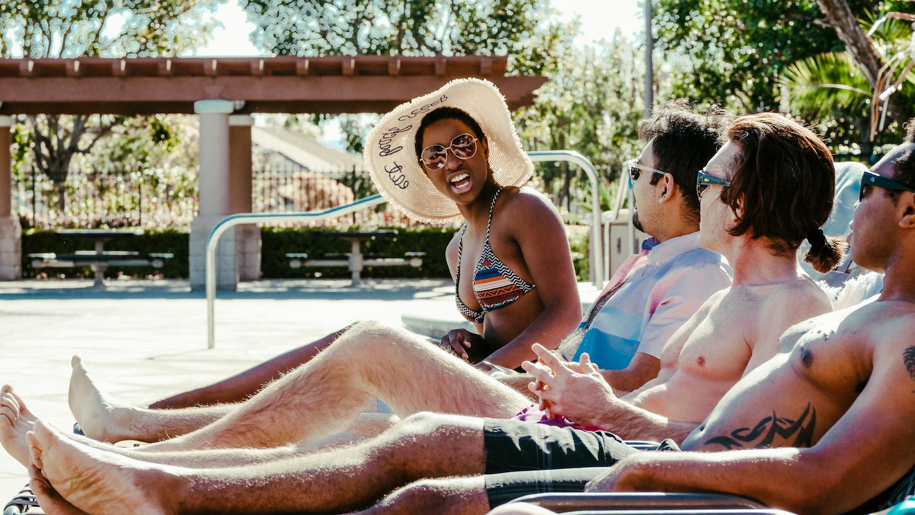 A group of friends enjoying a sunny day at a resort poolside, lying on sun loungers and wearing swimwear.