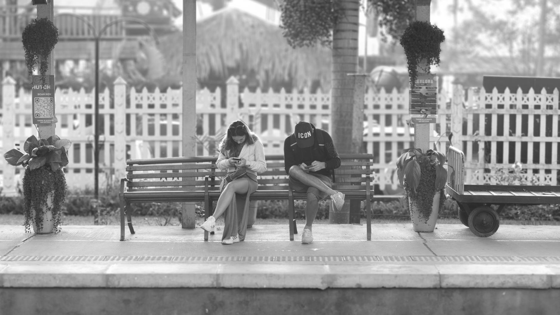 Two people seated at a Sri Lankan train station, engrossed in their phones.